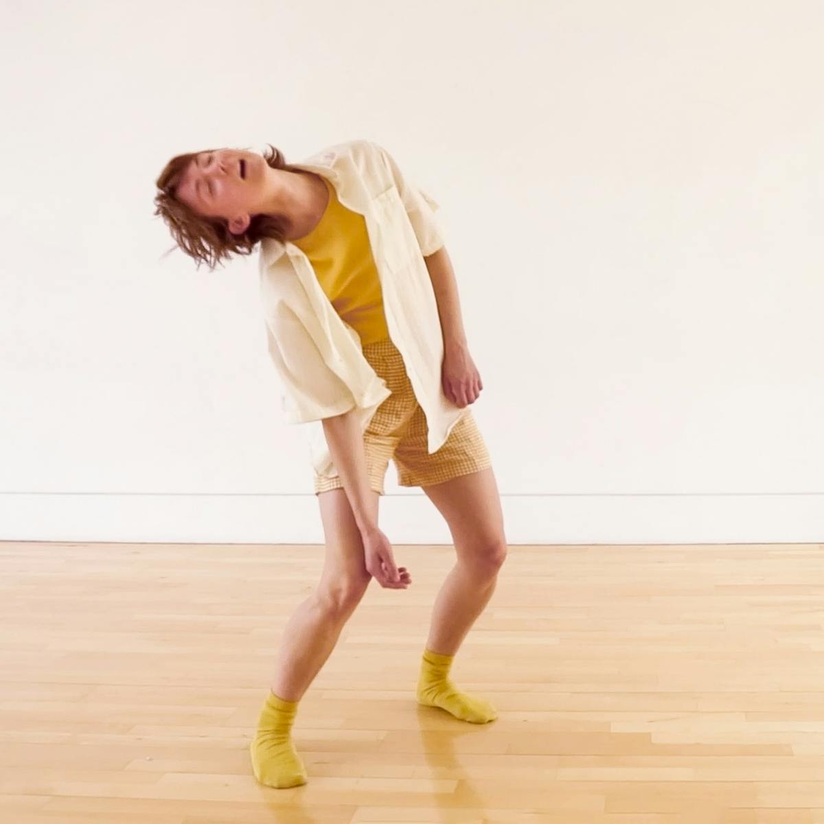 Photograph of a slim, white person in their mid twenties with short brown hair wearing yellow socks, shorts and a shirt. The figure is captured in movement, leaning to the left with bent knees and loose hanging arms. Their eyes are closed and their mouth is softly open. There is a plain white wall behind them and a smooth wooden floor beneath their feet. 