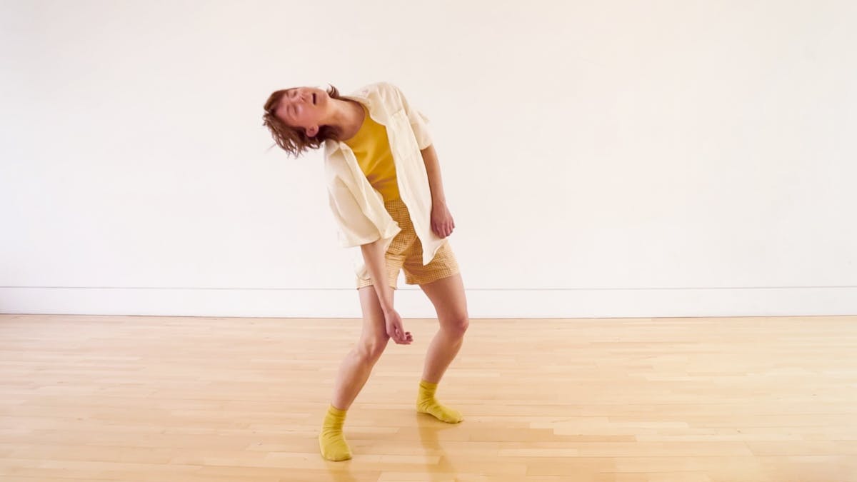Photograph of a slim, white person in their mid twenties with short brown hair wearing yellow socks, shorts and a shirt. The figure is captured in movement, leaning to the left with bent knees and loose hanging arms. Their eyes are closed and their mouth is softly open. There is a plain white wall behind them and a smooth wooden floor beneath their feet. 