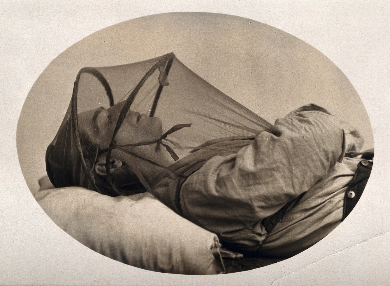 Black and white photograph of a sleeping man, showing his head and shoulders only. He is lying down, with his head resting on a pillow. He is wearing a mosquito net which covers his head and neck. It is tied on with a fastening beneath his chin.