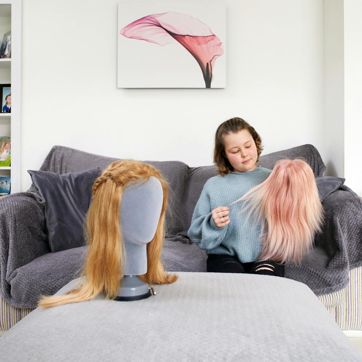 Photograph of a young girl in her living room, sat on a sofa. She is wearing a knitted grey jumper and ripped black jeans. She is holding a straight haired pink wig in her left hand and stretching out a few strands of hair with her right hand. She is looking down towards the wig with a slight smile on her face. In front of her is a foot stool, on which is another wig, this time blonde with a plait. The wig is supported in a mannequin head. Behind the young girl is a wood burning stove, a canvas artwork of a flower and a bookshelf containing DVD and family photographs in frames.