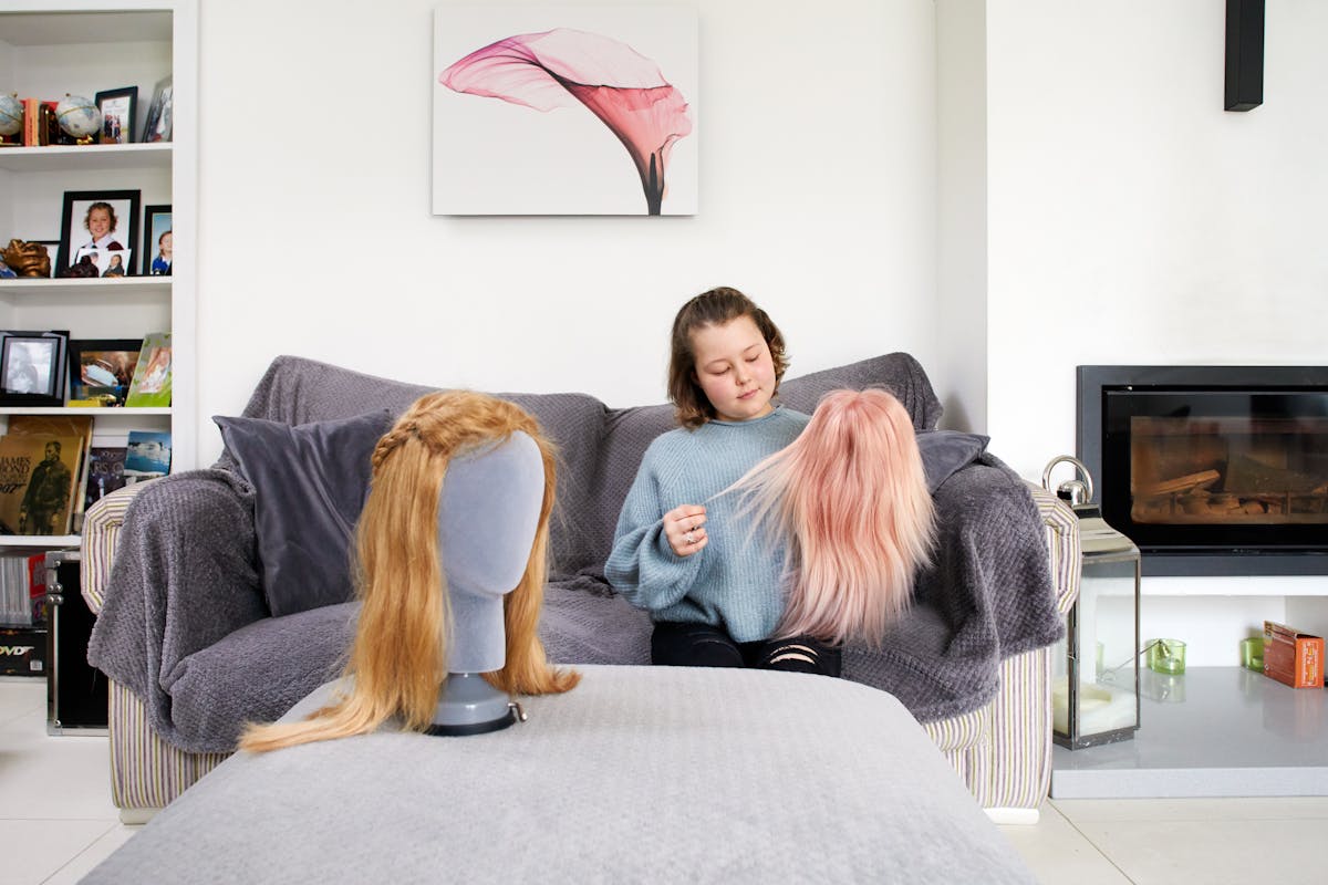 Photograph of a young girl in her living room, sat on a sofa. She is wearing a knitted grey jumper and ripped black jeans. She is holding a straight haired pink wig in her left hand and stretching out a few strands of hair with her right hand. She is looking down towards the wig with a slight smile on her face. In front of her is a foot stool, on which is another wig, this time blonde with a plait. The wig is supported in a mannequin head. Behind the young girl is a wood burning stove, a canvas artwork of a flower and a bookshelf containing DVD and family photographs in frames.