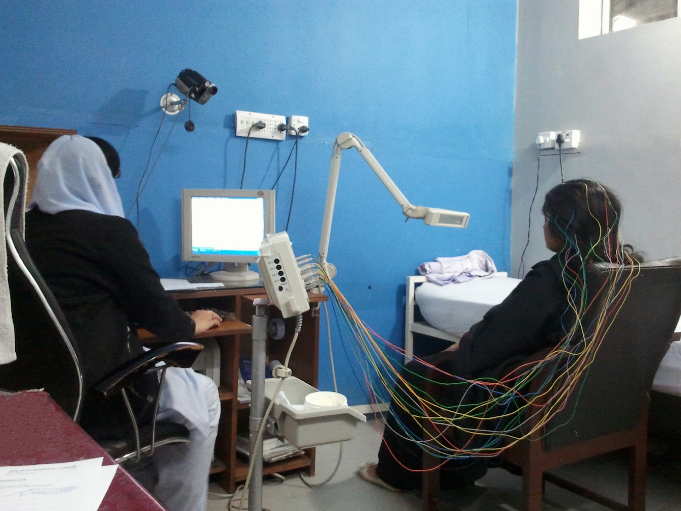 A woman seated with her back to the camera with wires in her hair having an EEG asa nurse watches a monitor next to her.