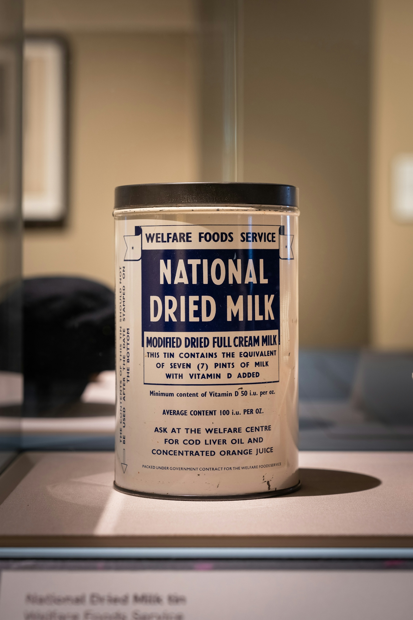 Photograph of a tin of dried milk from the 1950s, with the brand name, 'Welfare Foods Service, National Dried milk'. The tin is displayed in a perspex display case.