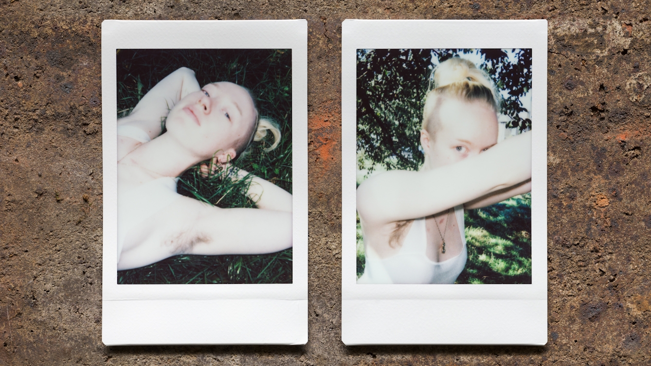 Photograph of two Instax Mini instant film prints in a line, resting on a textured brick surface. The two prints feature the same woman. The print on the left shows her lying on the grass, with her arms raised behind their head to show her armpit hair. The print on the right shows the same woman standing up but leaning forwards, supported by her hands and arms. She is looking towards the camera and her armpit hair can be seen.