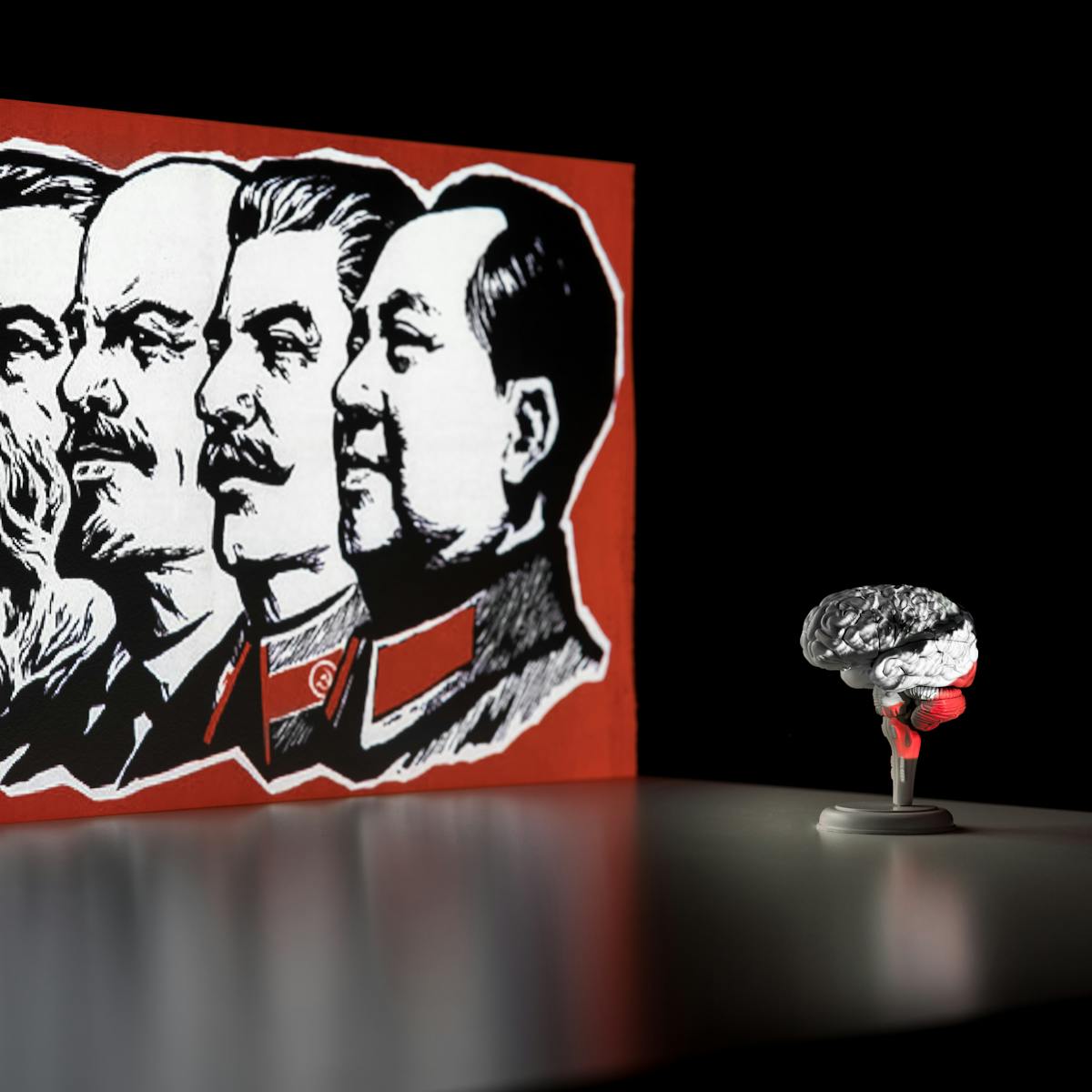 Photograph of a dark corner of a room. On the wall is a projected illustration of the heads of Marx, Engels, Lenin, Stalin and Mao Zedong, all in profile. The illustrated heads are monotone set within a bright red border. In front of the projected image is a flat tabletop which shows a slight reflection of the illustration. Standing on the table to the right is a small model of a brain, resting on a stand. Part of the projected image washes across the surface of the brain, covering it in the image and its colours.