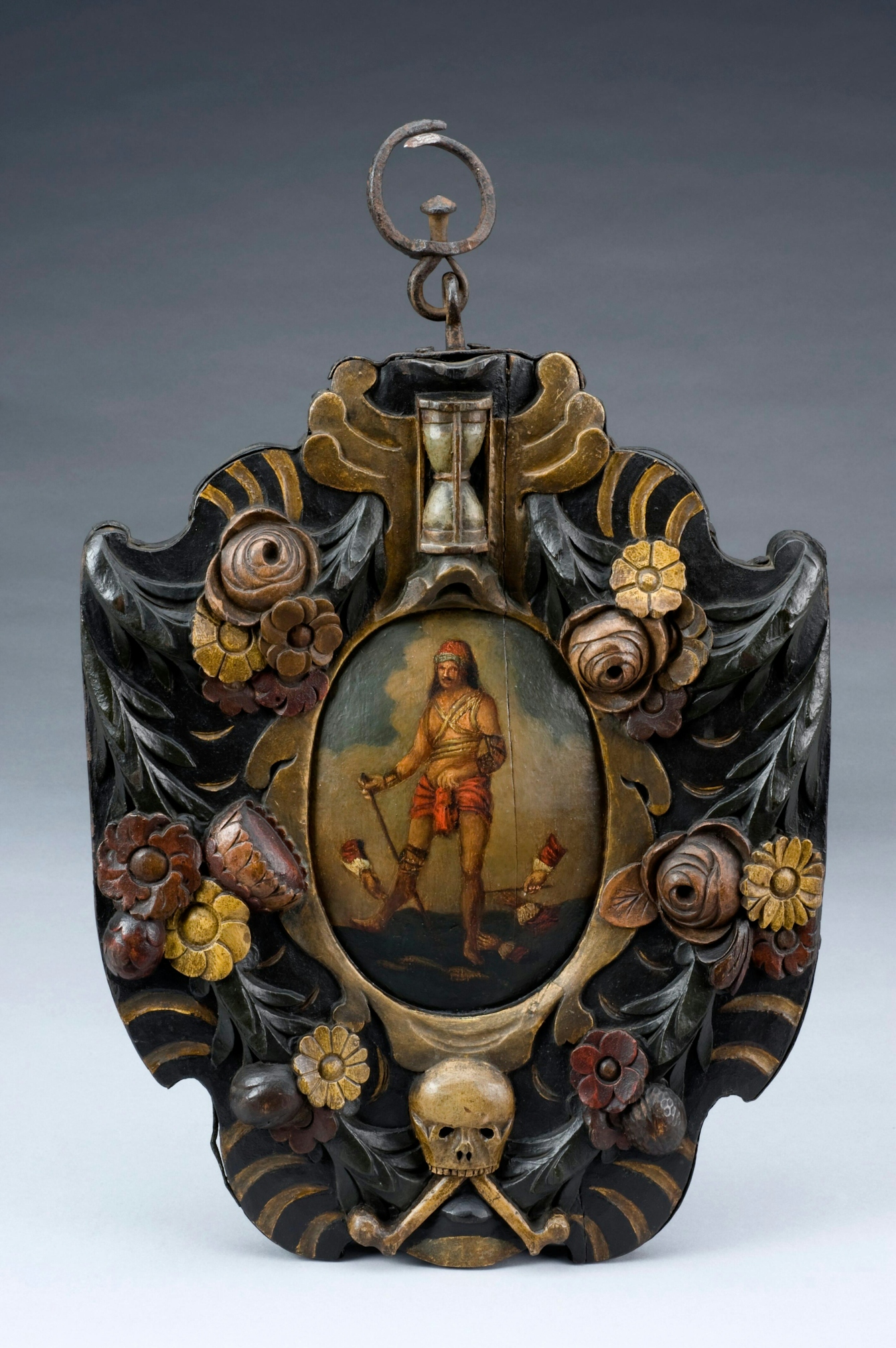 Colour photograph of a wooden carved and painted plaque. In the centre is a picture of a man holding a stick as three disembodied hands cut his legs with small blades. Around the frame are flowers, with a sand timer at the top and a skull at the bottom. 