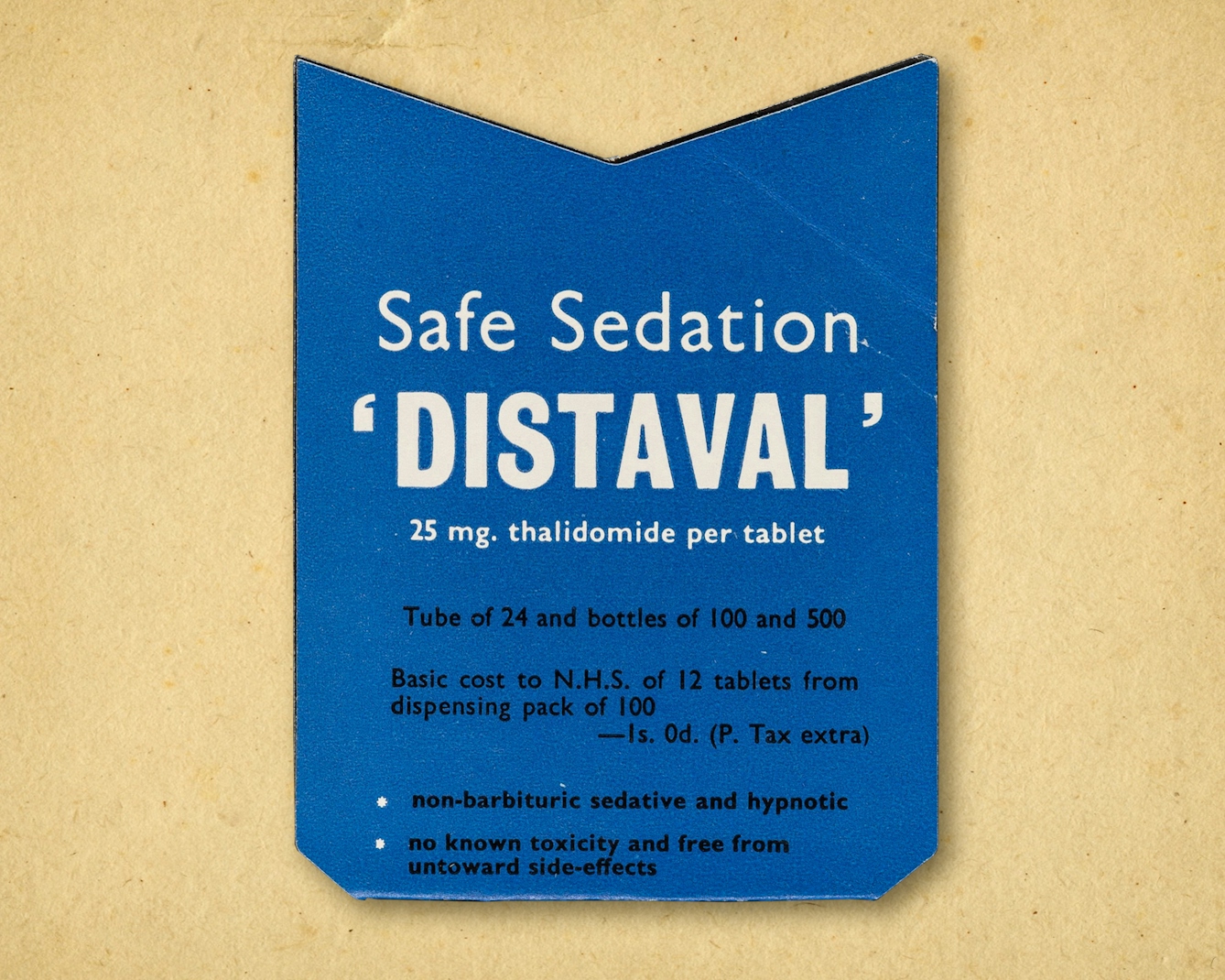 Photograph of a die-cut advertisement issued by The Distillers Company (Biochemicals) Limited for the sedatives Distaval and Distaval Forte, circa 1958, resting on a brown paper textured background. The advertisement is a rectangular blue shape with a 'v' cut out of the top. On the front in black and white text of varying sizes are the words, "Safe Sedation 'DISTAVAL' 25mg thalidomide per tablet. Tube of 24 and bottles of 100 and 500. Basic cost to NHS of 12 tablets from dispensing pack of 100 - 1s. 0d. (P. Tax extra). Non-barbituric sedative and hypnotic. No known toxicity and free from untoward side-effects."