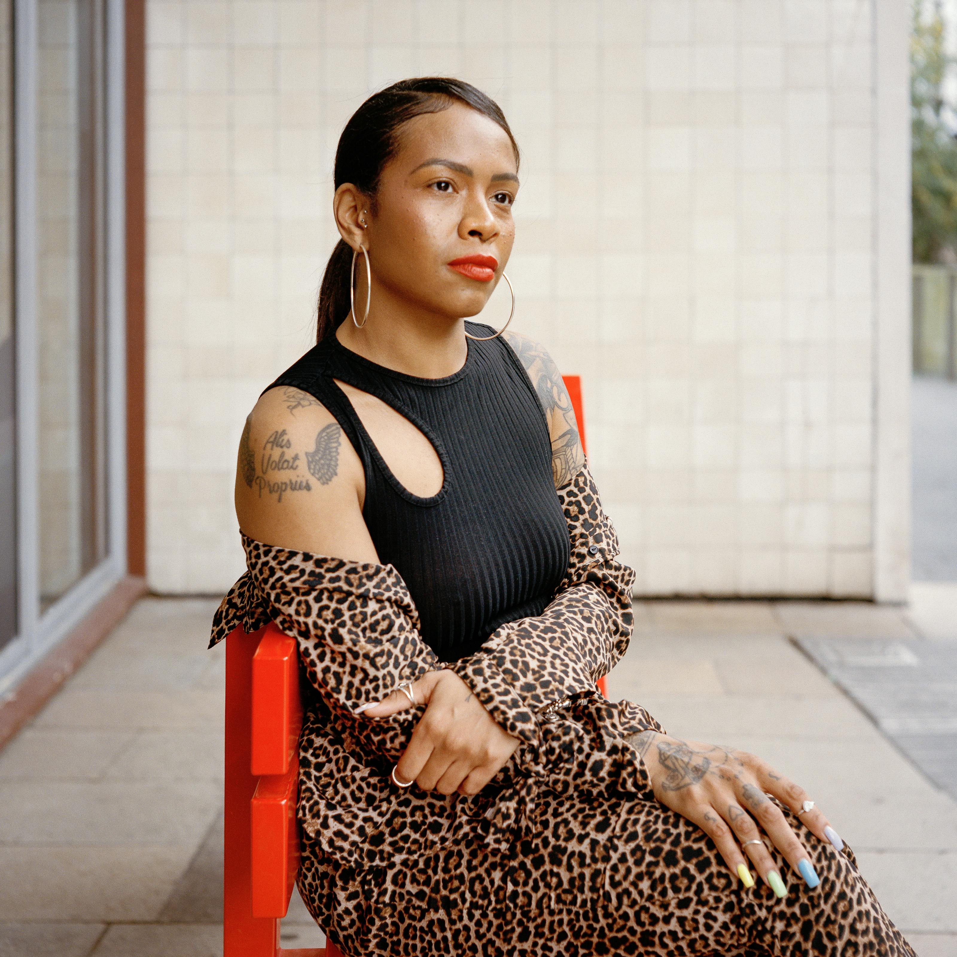 Portrait photo of Lauren,  a young woman with black hair, sitting on an orange bench outside. She has several tattoos on her arms and is wearing a black sleeveless top, leopard print shirt and a leopard print skirt. She has large hoop earrings and is wearing an orange red lipstick. Her nails are painted in a variety of colours.

She is looking into the distance, slightly smiling. Behind her, there is a building and a tiled wall. 