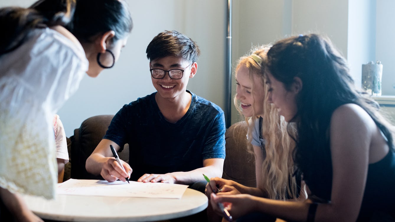 Four young people sitting around a table with pens and one large piece of paper that they're writing on collaboratively, they are looking at the paper and smiling.