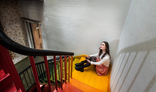 Photograph of Emily Bashforth sitting on stairs which are painted in a variety of bright colours. The stairs have a red handrail. 

Emily is looking up, towards the top left of the photograph. She is wearing a cream jumper, orange plaid skirt, black boots and red lipstick. She has long brown wavy hair. 

To the left, there is some small text superimposed upon a grey wall. Beneath the text is a wooden doorway and wooden floorboards. To the left of the door is a wall with a floral patterned wallpaper. 
