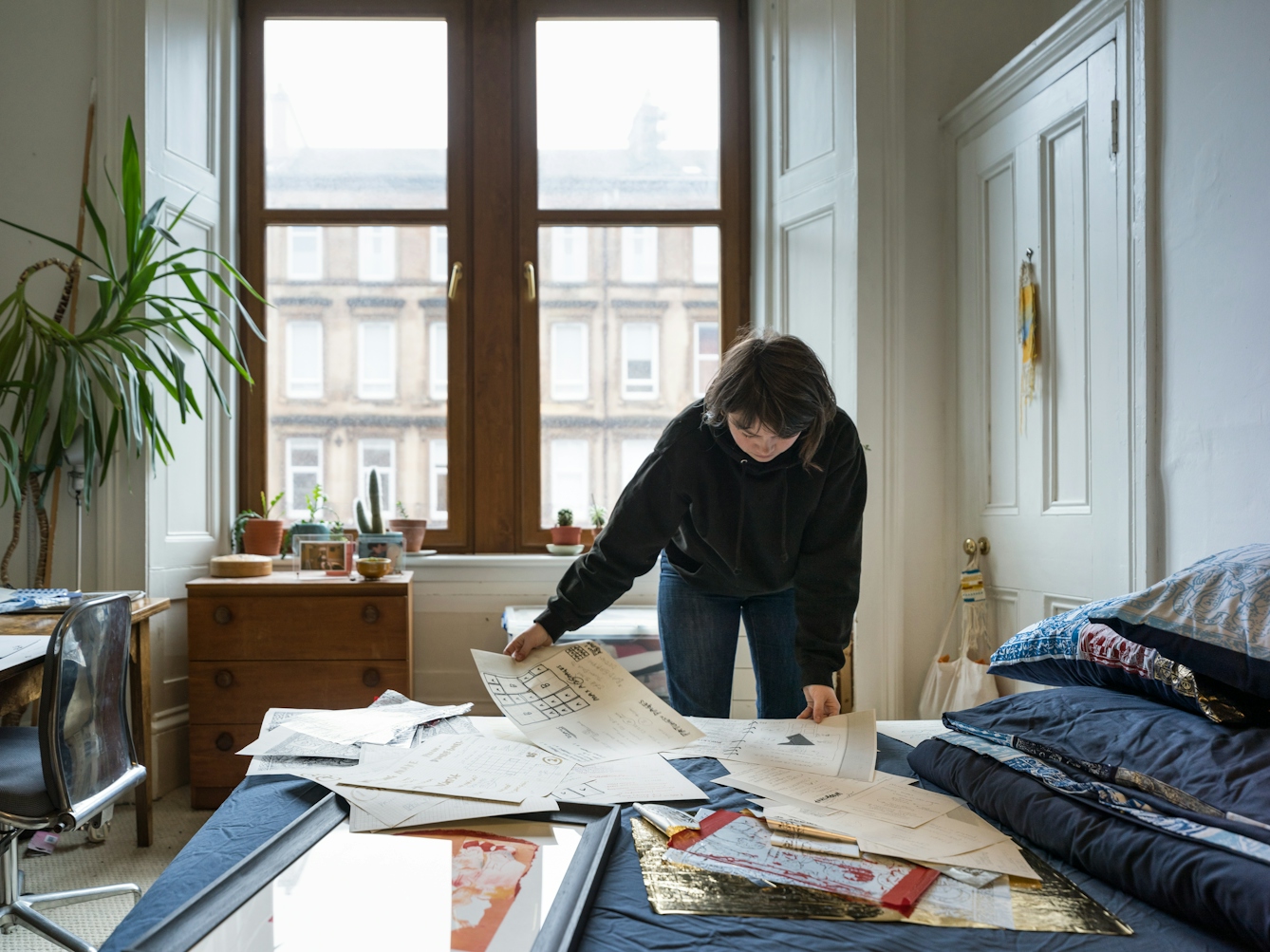 Photograph of a young woman bending over her bed holding sheets of paper in her hands. The bed is covered in more sheets of paper containing drawings and sketches. Behind her are large windows, a chest of drawers, a pot plant and the corner of a desk.