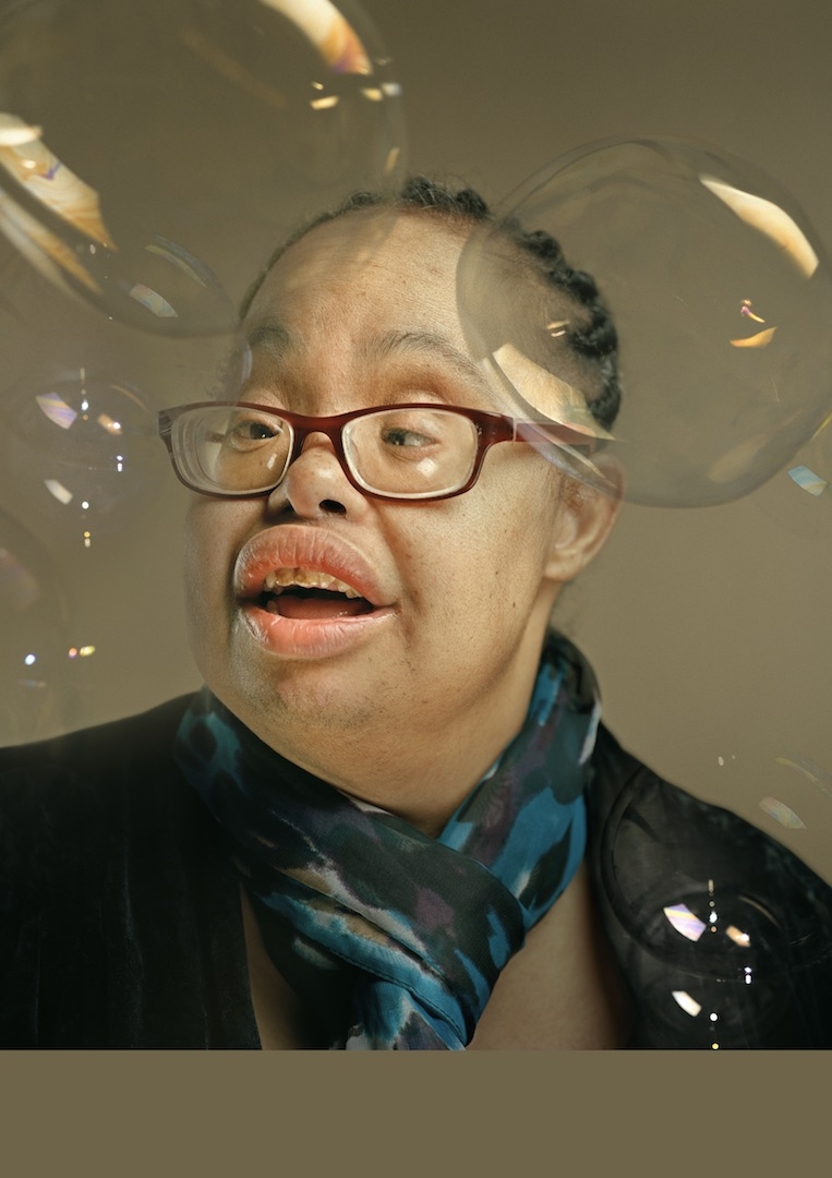 Colour photograph portrait of the head and shoulders of Cheryl Lyte, a woman with braided hair wearing glasses and a scarf. There are bubbles surrounding her face.