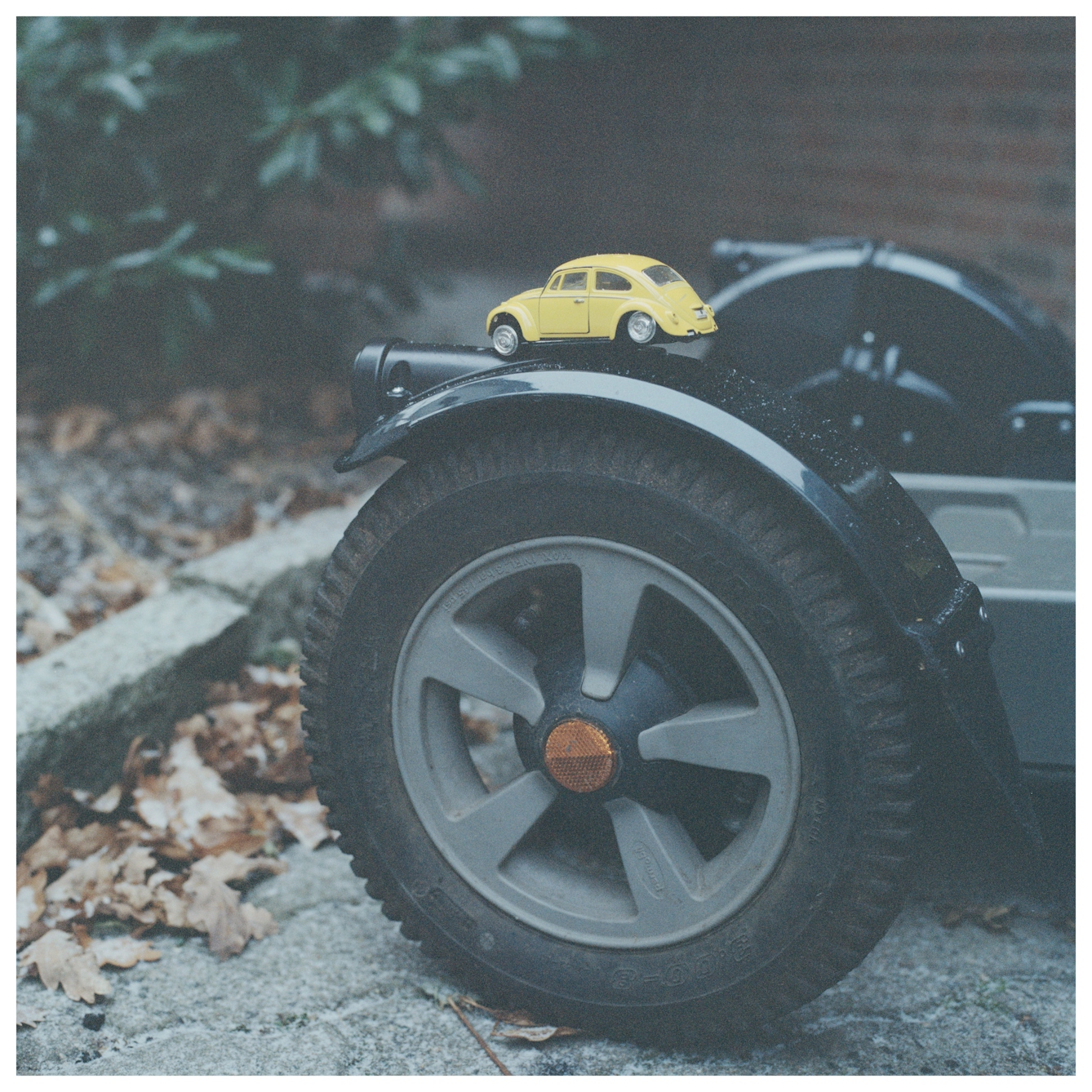 Photograph of the front left wheel of an off road electric wheelchair. The wheel almost fills the frame with a paved garden scene behind. Resting on top of the wheel's mudguard is a toy yellow VW beetle car. The tyres have been lost leaving just the bare silver hubs.