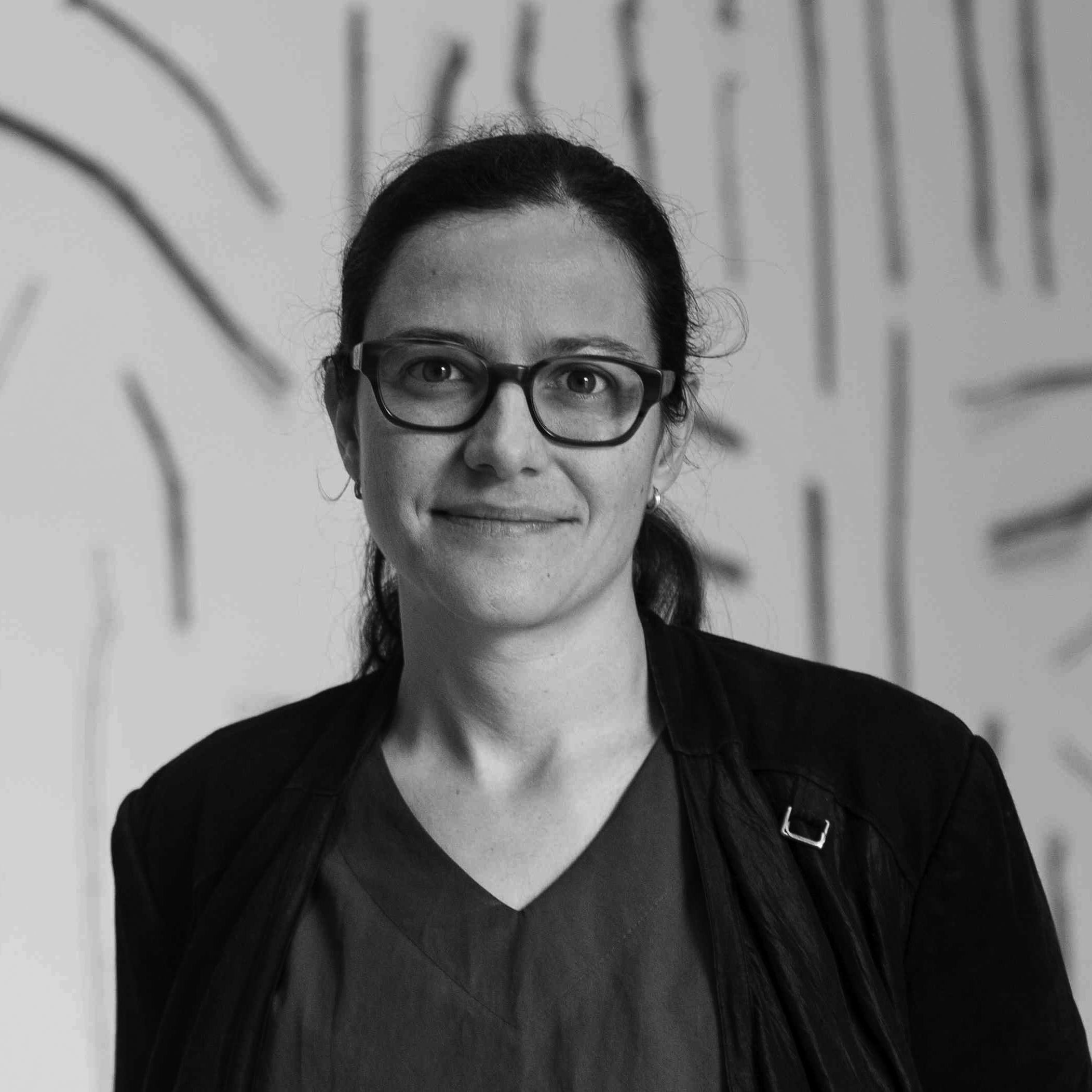 Black and white, head and shoulder photograph of a young woman. She has her hair tied back and is wearing glasses. She is standing in front of a blurred background with black lines.