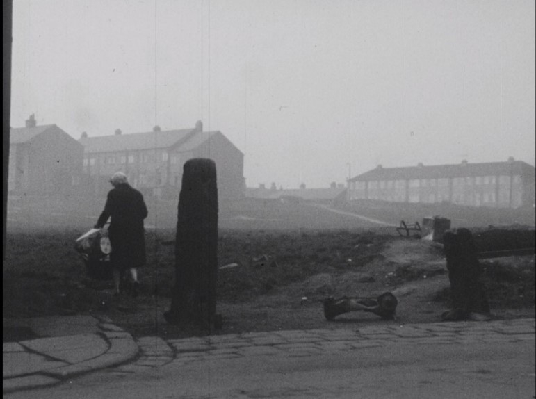 Black and white film-still showing a foggy scene of rows of houses. A woman with her back to us, wearing a dark coat and a headscarf, pushes a pram away from the camera and towards the houses. In the foreground, there appears to be debris and rubbish. 