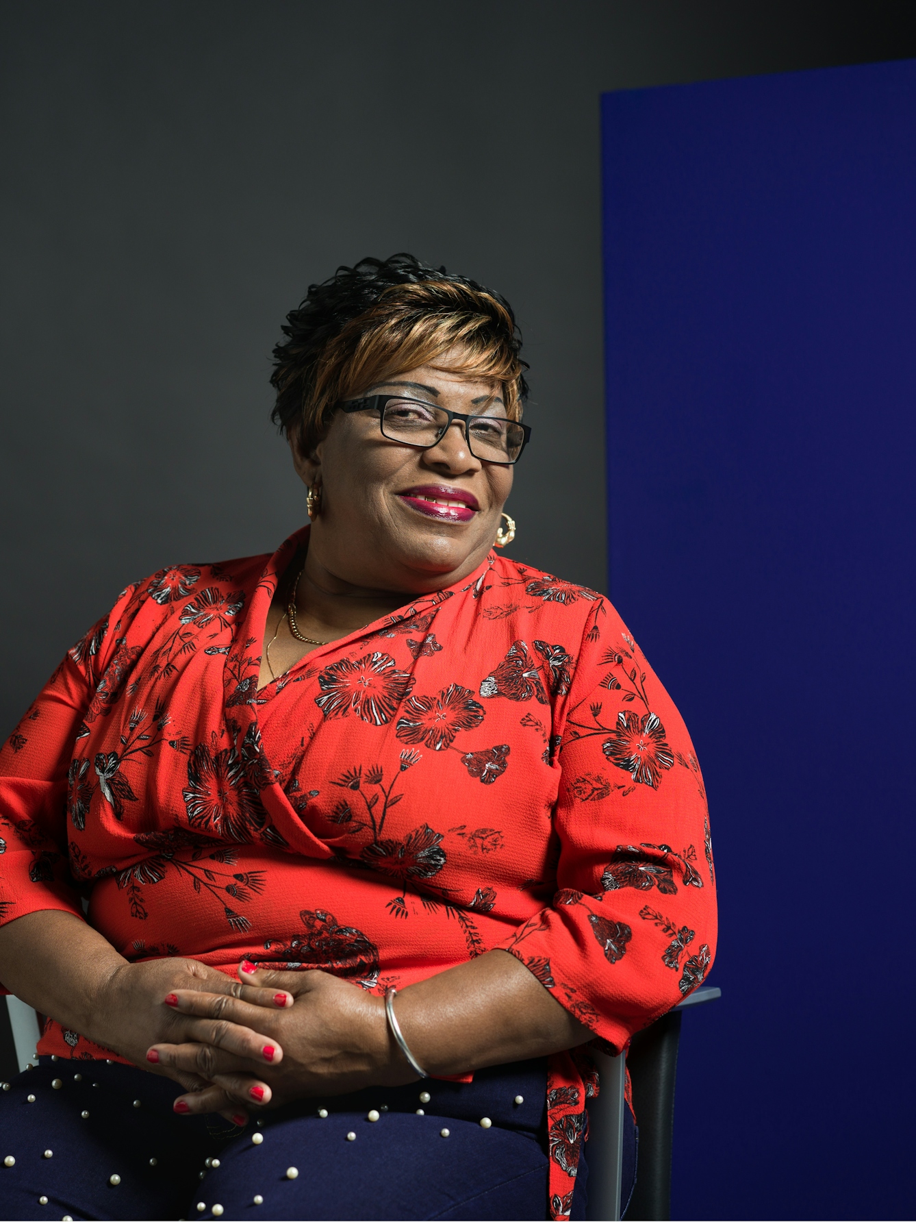 Colour photograph half-length portrait of Joyce Williams, a black woman sitting in a chair. Joyce is smiling and has short hair with a fringe, red lipstick and gold hoop earrings. She wears a floral red shirt, with matching red nail polish on her fingers, and blue trousers with white polka dots. 
