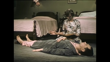 A man in pyjamas and a dressing gown lies down on the bedroom floor with a woman kneeling next to him and reading from a book