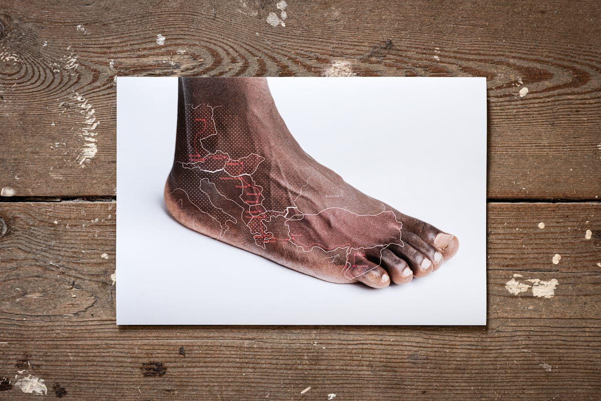 Photograph of a photographic print on a weathered, paint spattered wooden floor. The print shows a naked human foot on a white background. Superimposed on top of the foot is a simple line map in white and red, showing the outline of Italy and the eastern mediterranean shoreline. 