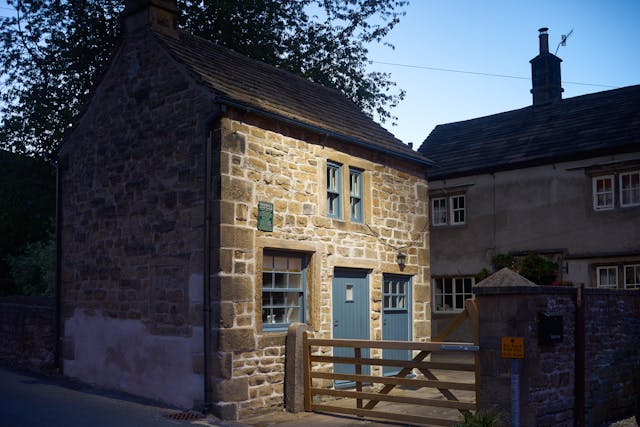 Photograph of a stone built house with 5 bar gate, shot at dusk. On the wall of the house is a green plaque detailing those how died in the house during the plague.