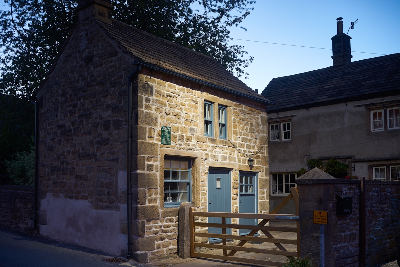 Photograph of a stone built house with 5 bar gate, shot at dusk. On the wall of the house is a green plaque detailing those how died in the house during the plague.