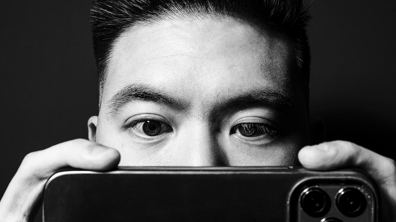 Black and white photographic portrait of a close-up of a man's face. He is holding up a smartphone close to his eyes such that it obscures his nose and mouth. His eyes are directed towards the phone's screen. His finders are visible holding the phone on either side and the camera lenses of the phone appear in the bottom right corner of the image. The man's face and hands are spotlit in a small circle of light. He is standing against a black background which means he is surrounded by darkness.