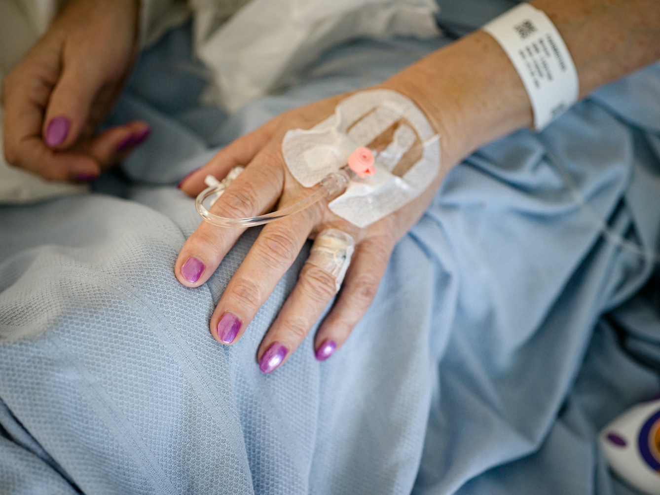 Photograph of the hands of a patient with glittery purple painted fingernails, resting on a light blue hospital bedsheet. The patient's hospital name tag and cannula can be seen.