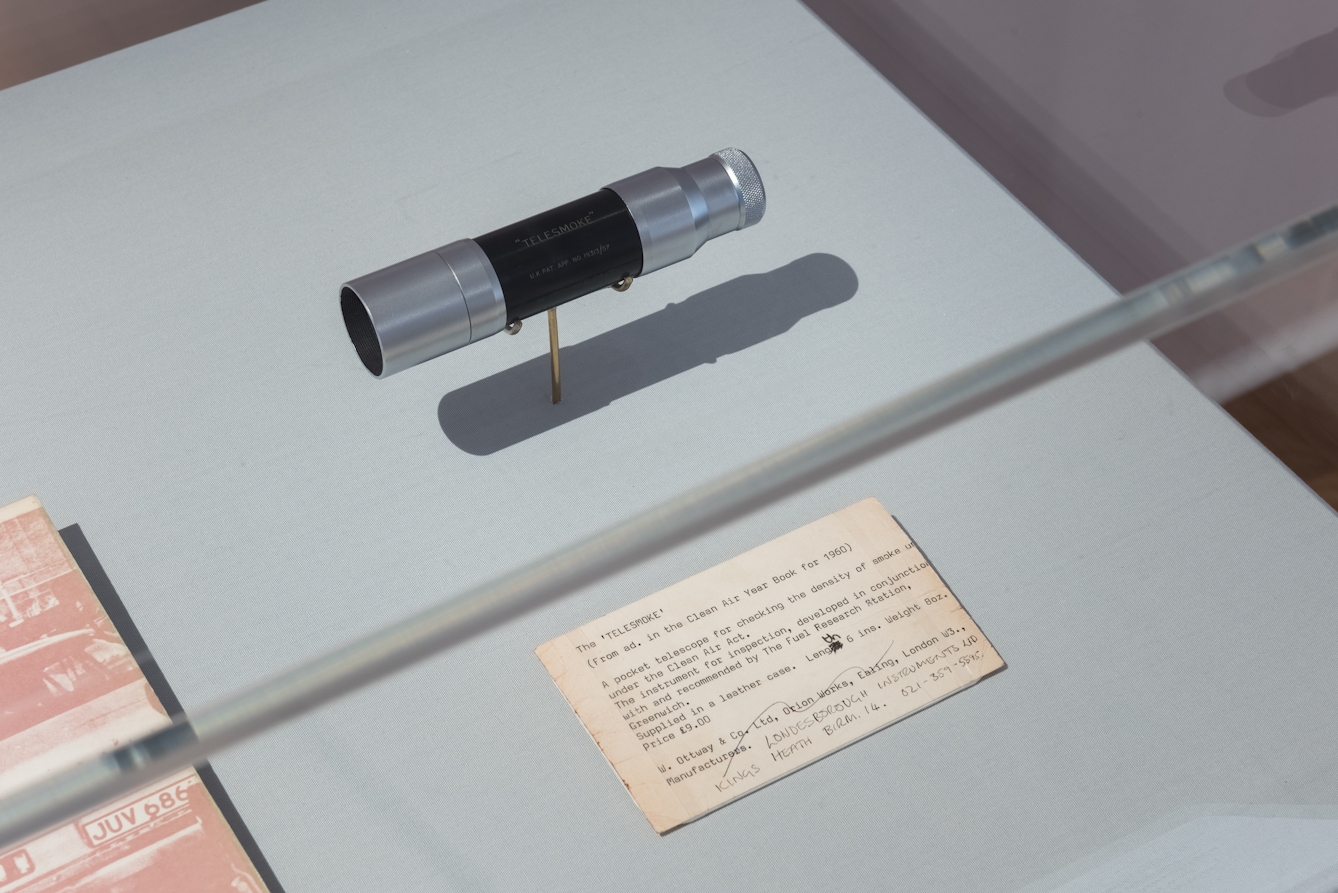 Colour photograph of a small metal telescope elevated on a stand. There is a piece of paper with text next to the telescope, which explains what it is used for. Both the telescope and piece of paper are inside of a glass box. 