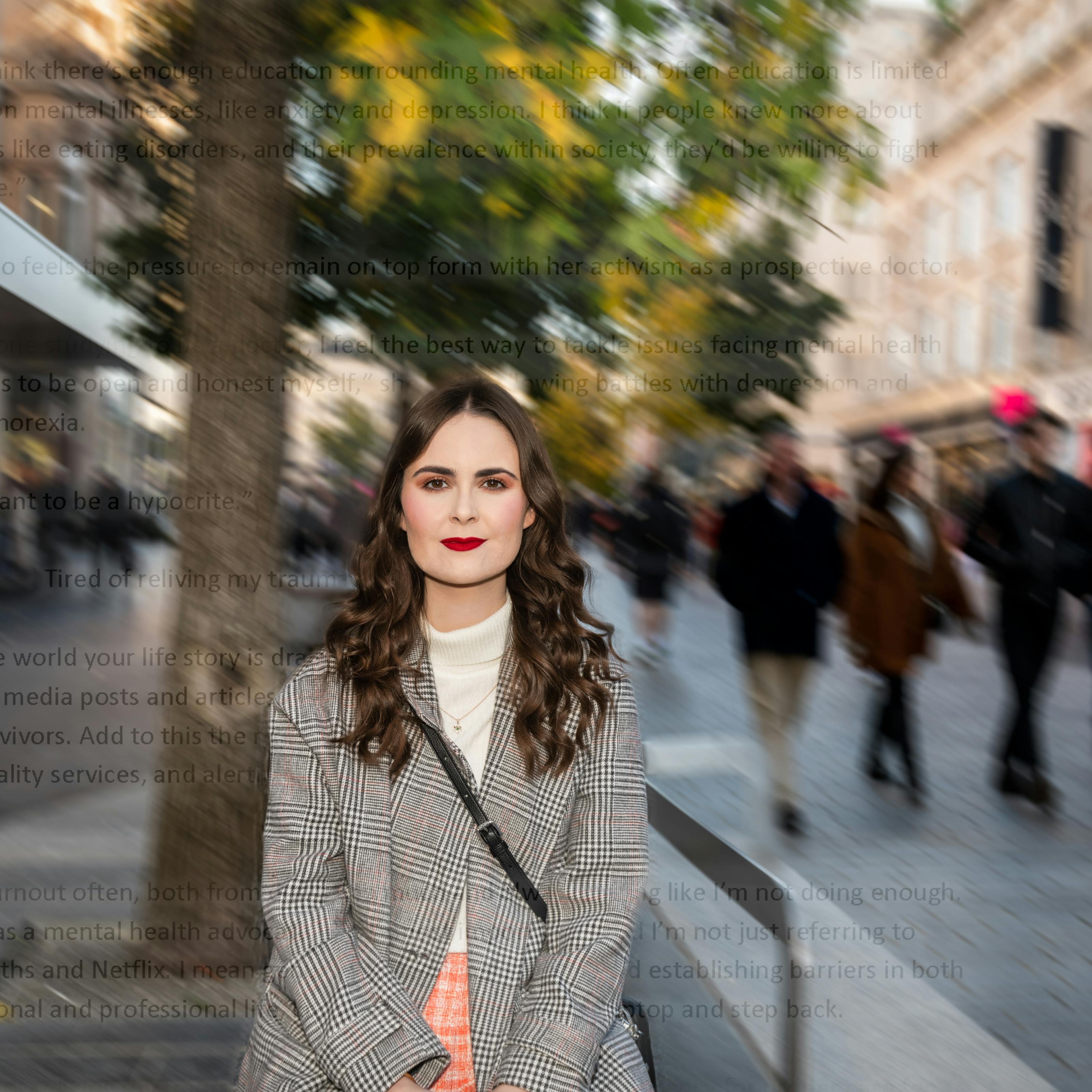 Portrait of Emily Bashforth. She is standing on a busy street which is blurred in the background. She is wearing a cream jumper, a checked coat, orange plaid skirt and red lipstick. She has long wavy brown hair and is looking directly at the camera. 

Superimposed on the image are lines of text - mostly unreadable. A visible phrase reads 'Tired of reliving my trauma'. 
