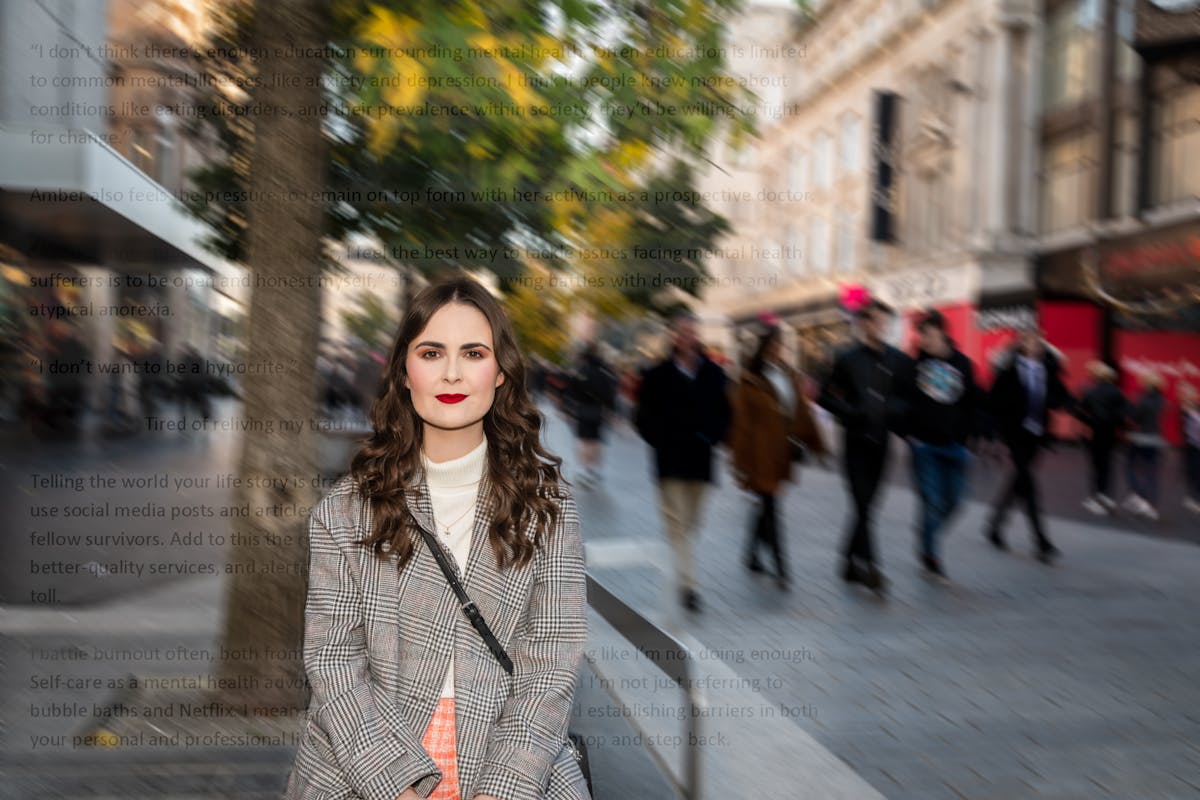Portrait of Emily Bashforth. She is standing on a busy street which is blurred in the background. She is wearing a cream jumper, a checked coat, orange plaid skirt and red lipstick. She has long wavy brown hair and is looking directly at the camera. 

Superimposed on the image are lines of text - mostly unreadable. A visible phrase reads 'Tired of reliving my trauma'. 