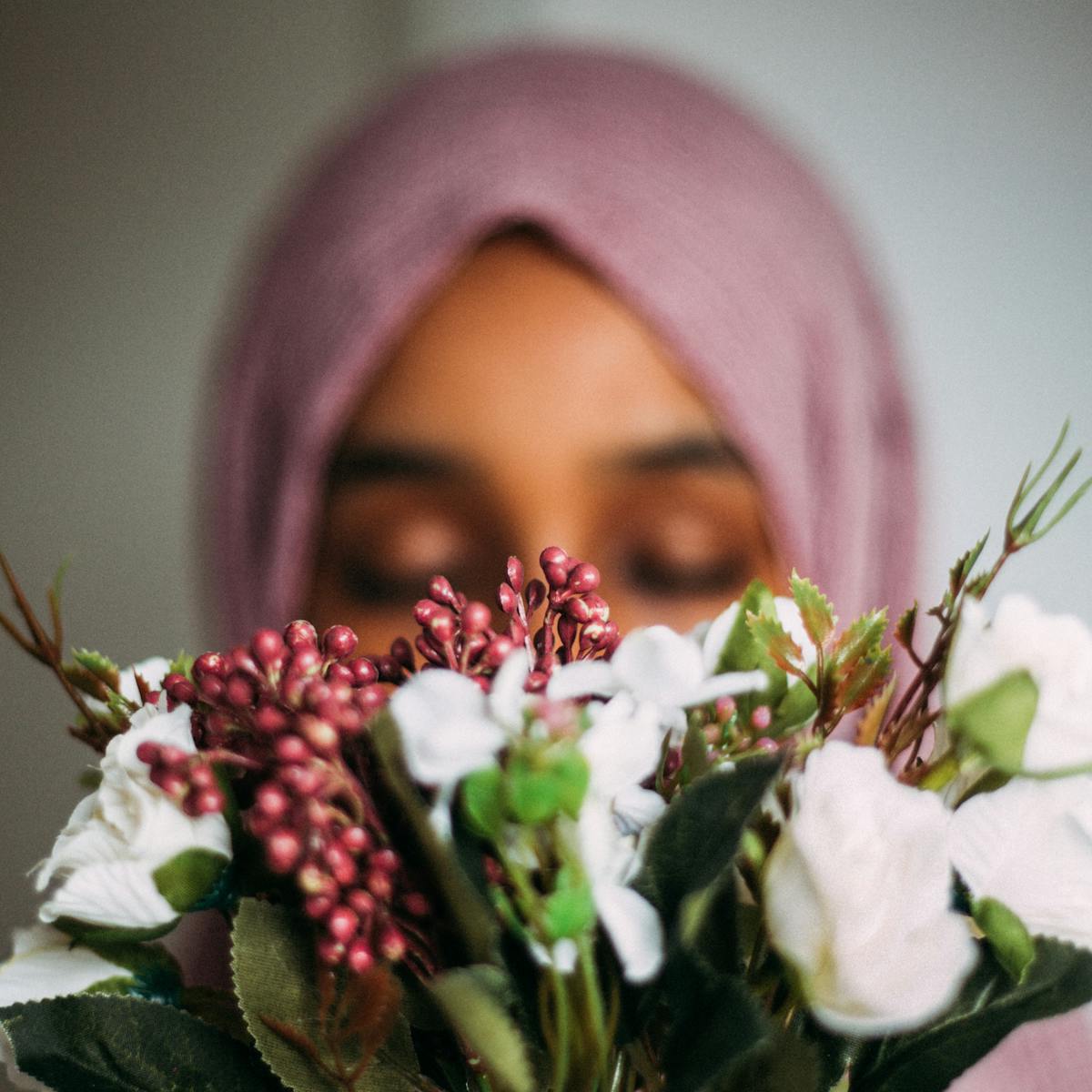 Photograph of a woman wearing a pink head scarf facing towards the camera. Her face is out of focus and partially obscured by a bunch of flowers.