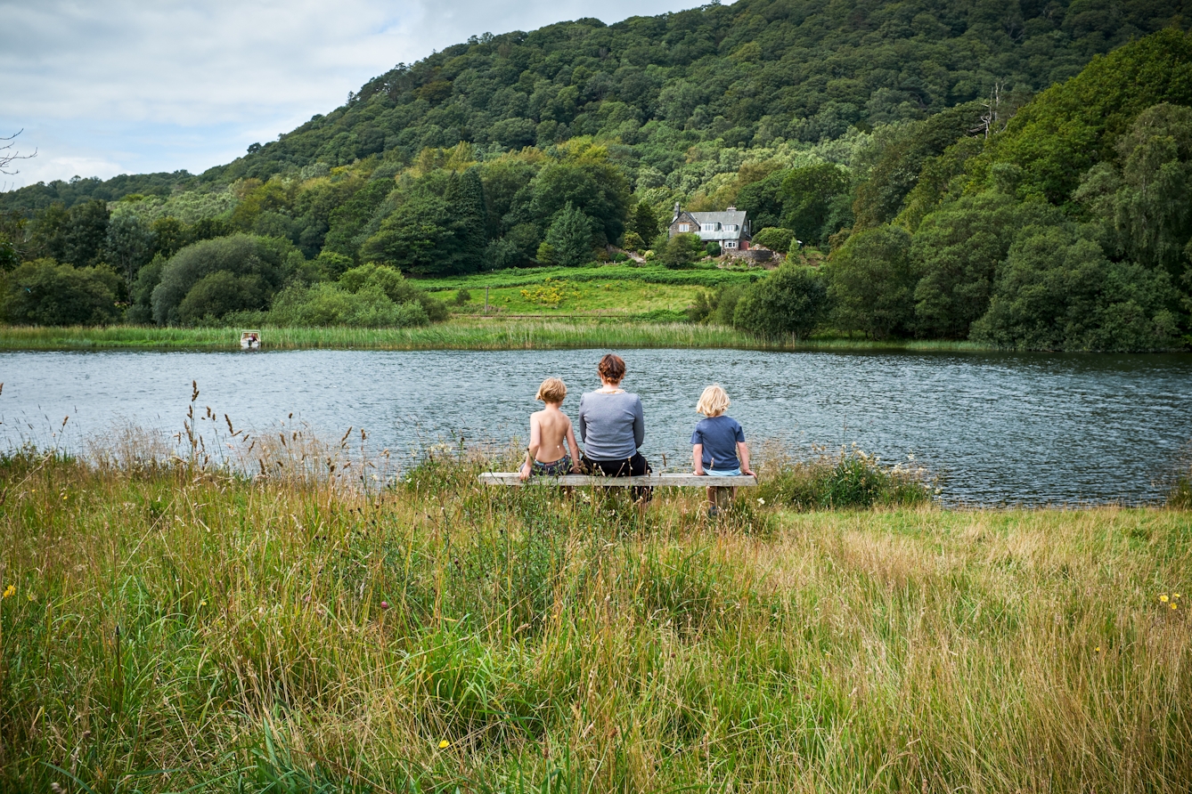 Photograph of an adult and two children sitting in a field, on a wooden bench which is facing a lake. Across from the lake there is a woodland and a small house. 

The adult has short brown hair and is wearing a long-sleeve grey t-shirt. Sat on their left is a child with blonde hair who is wearing colourful shorts. On her right is another child with light blonde hair and a dark blue t-shirt. 