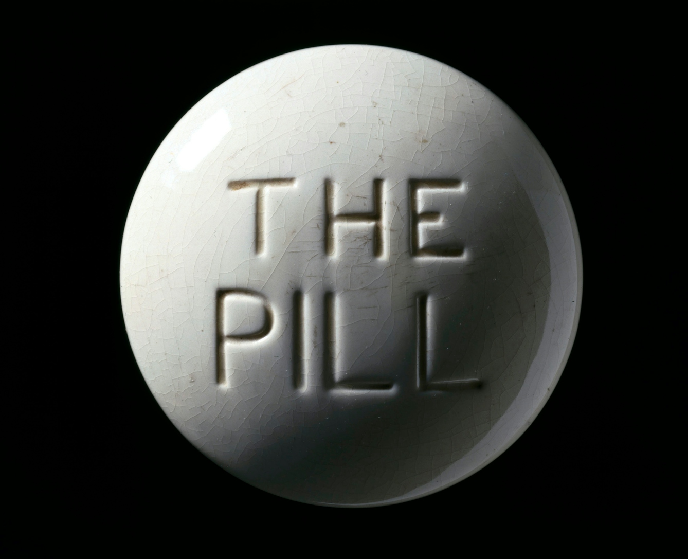 Photograph of a contraceptive pill model, Europe, c. 1970. White crackled glaze, with debossed "THE PILL" text in centre, on a black background.