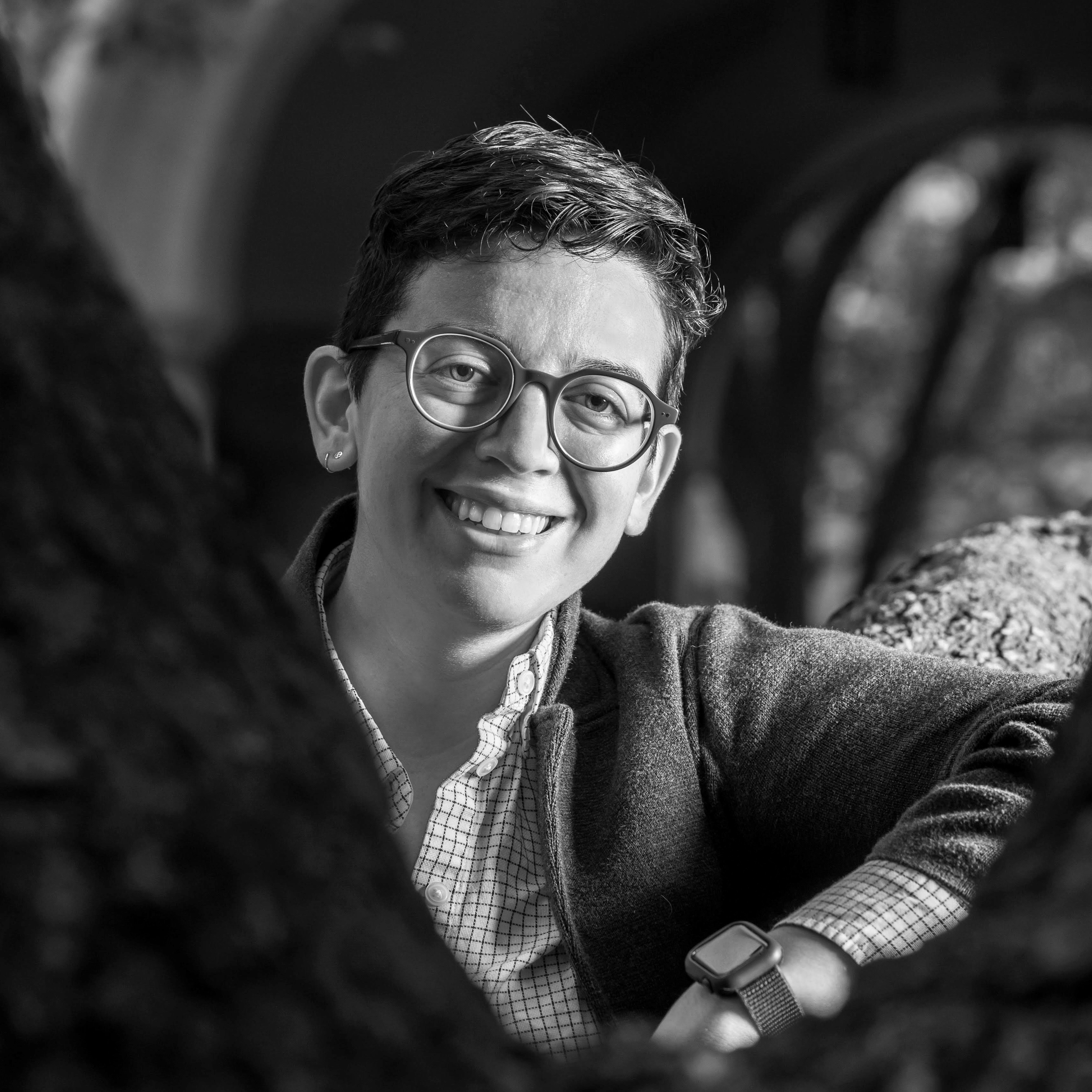 Black and white photograph of Carly B. Boxer, a person with short brown hair and glasses wearing a checkered shirt and grey jacket, with blurry stone archways in the background.