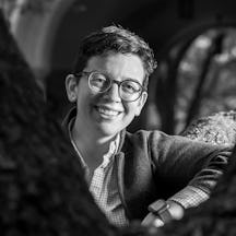 Black and white photograph of Carly B. Boxer, a person with short brown hair and glasses wearing a checkered shirt and grey jacket, with blurry stone archways in the background.