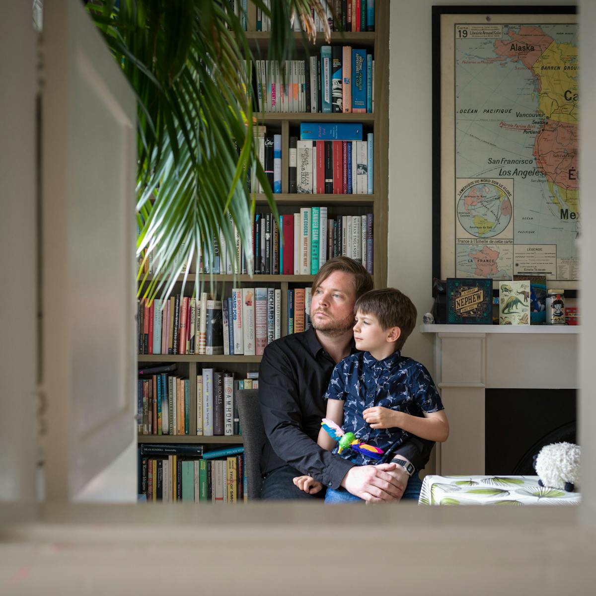 Photograph of a man sitting in a dining room with his arms around his son, sitting on his lap. Behind them is a bookcase and part of a world map. They are framed by the opening of a hatchway in the wall.