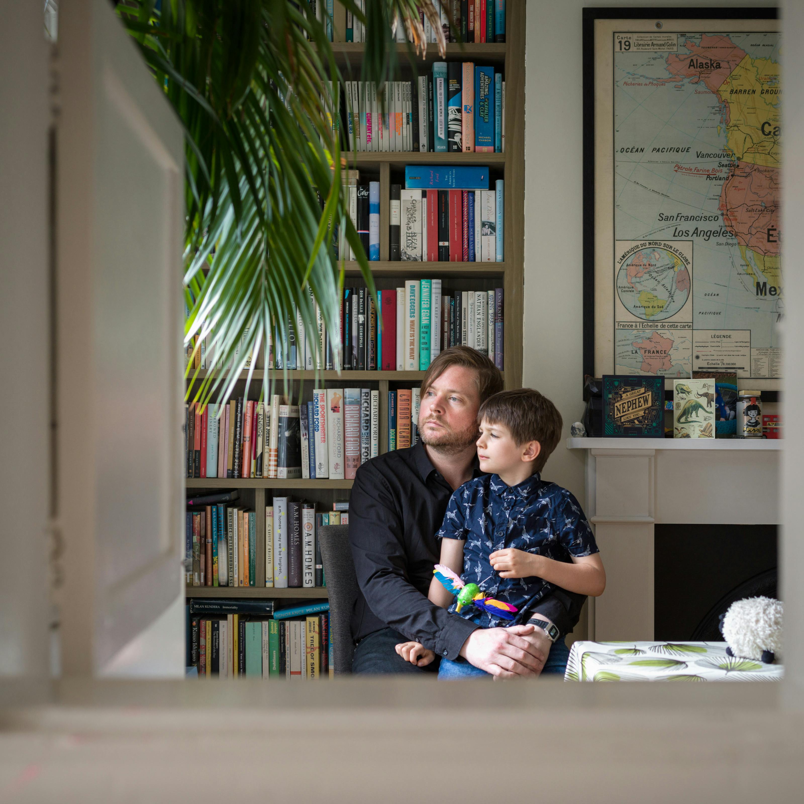 Photograph of a man sitting in a dining room with his arms around his son, sitting on his lap. Behind them is a bookcase and part of a world map. They are framed by the opening of a hatchway in the wall.