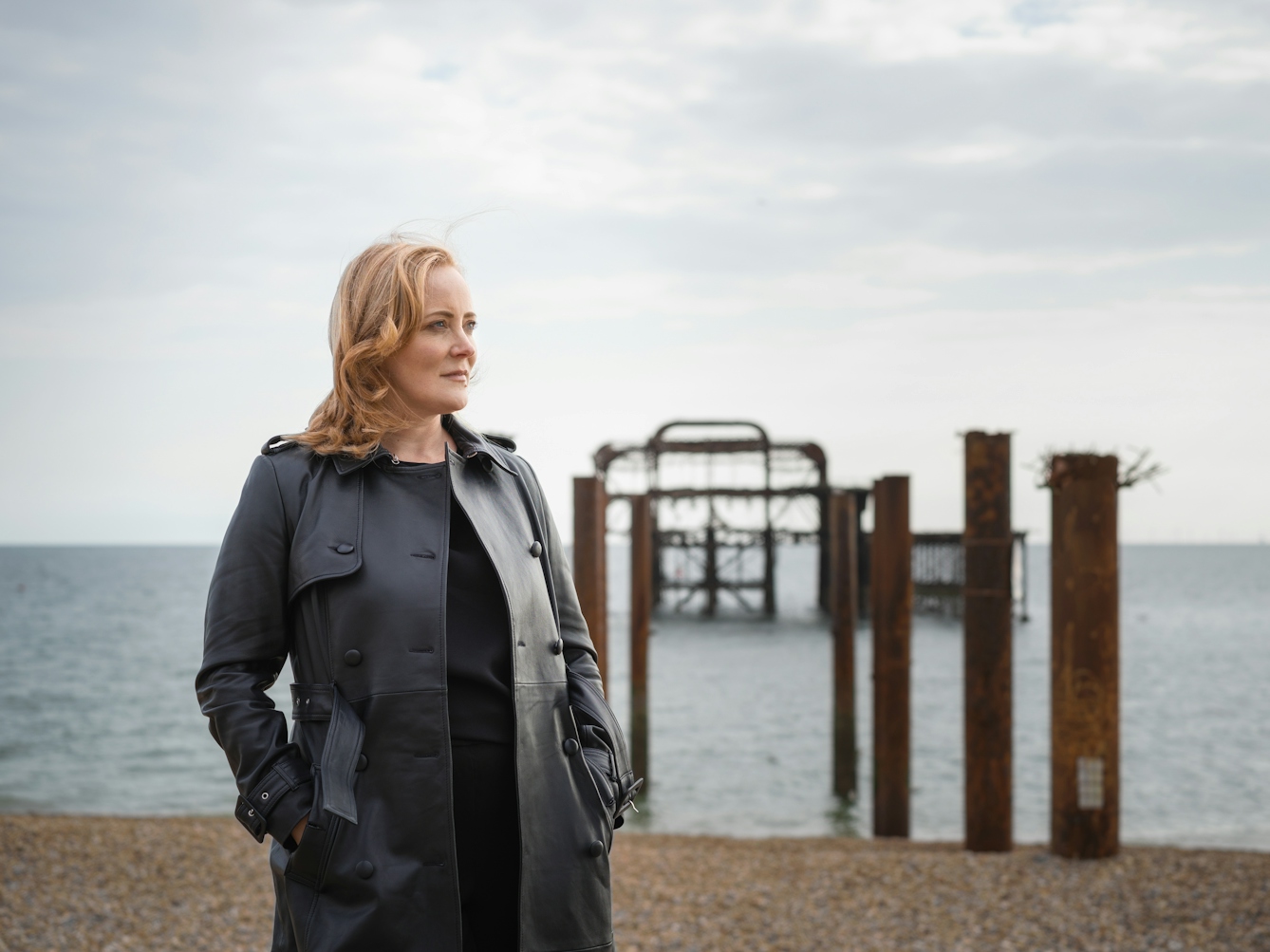 Photograph of a blonde woman as she gazes to the right of the frame on a sea front.  Out of focus behind her is a pebble beach with a burnt out pier.
