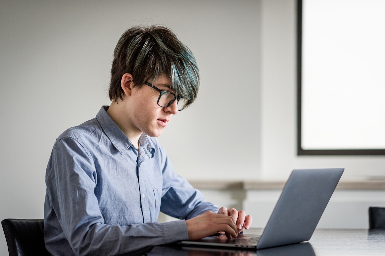 Photograph of a young man sat in white walled room at a black glossy table. He is looking at a laptop computer screen his hands poised over the keyboard. In the background is the corner of a window, from which light is flooding in.