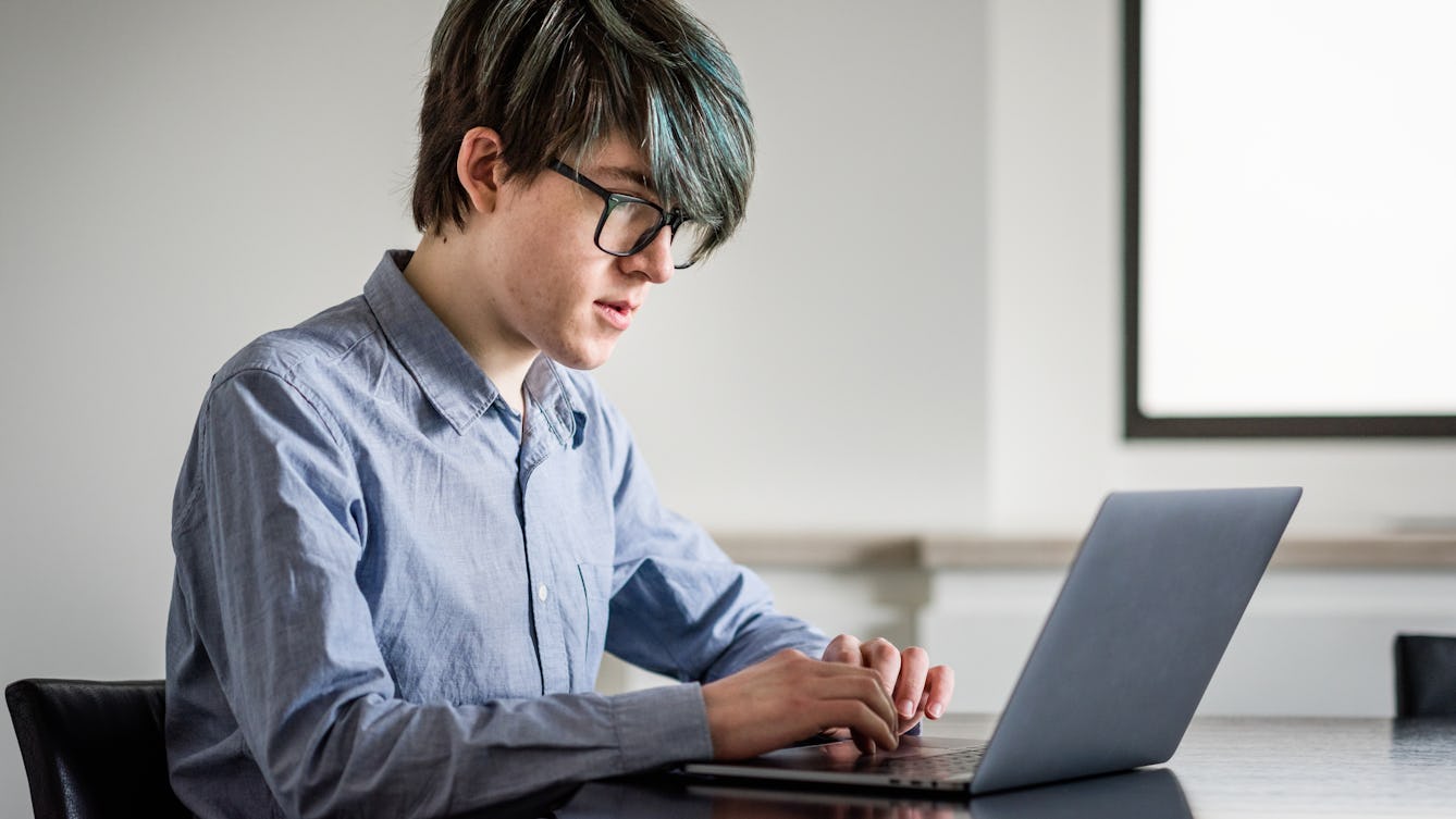 Photograph of a young man sat in white walled room at a black glossy table. He is looking at a laptop computer screen his hands poised over the keyboard. In the background is the corner of a window, from which light is flooding in.