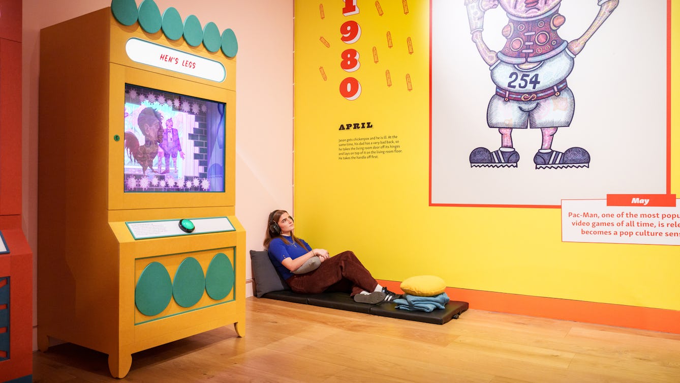 A woman sits on a mat in the corner of a room with yellow walls. She is wearing headphones, holding a cushion, and looking upwards.