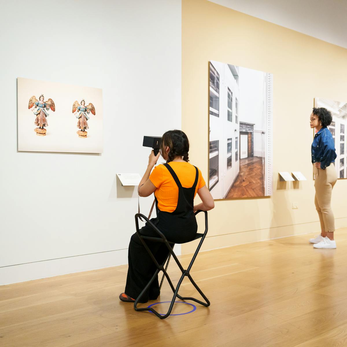 Photograph of a gallery exhibition space showing large photographic prints mounted on the walls and free standing display boards. The colours of the room are light whites and yellows. Three people are exploring the space. One visitor wearing an orange t-shirt, sits on a gallery folding chair holding a stereoscopic viewer to their eyes, focusing on a photographic print on the wall depicting a ceramic angel figure, shown in duplicate. Another visitor to the right wearing a green jumper is also looking through a stereoscopic viewer at a print on the wall. In-between them another visitor in a denim shirt is looking at a large photographic print of a museum storeroom.