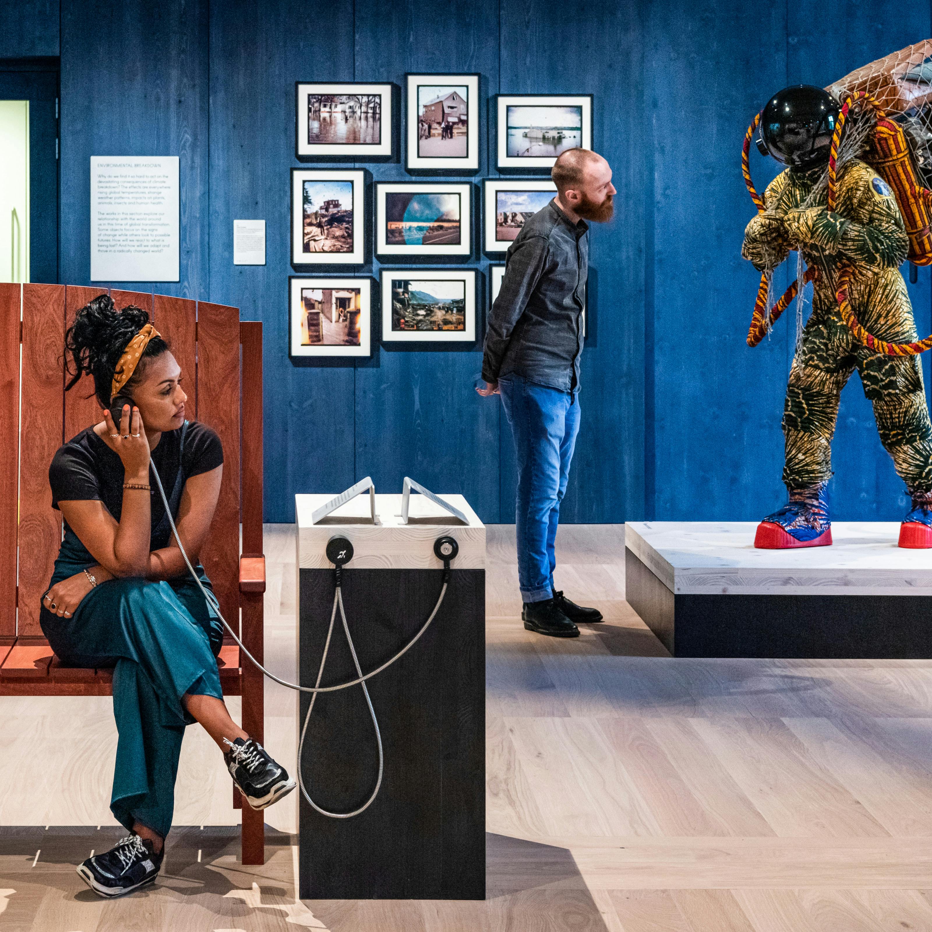 Photograph of an exhibition gallery space, with a blue stained wood wall in the background, in front of which a young man looks at a life-size artwork of a figure resembling an astronaut. In the foreground a young woman sits on a wooden bench holding an audio speaker to her ear.