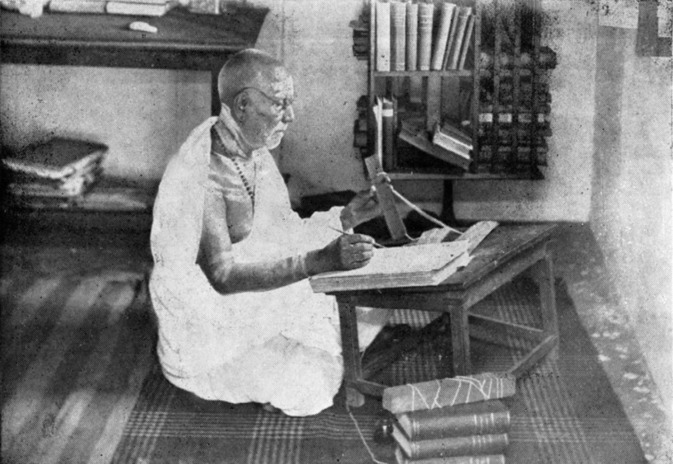 An old indian man (possibly a Hindu priest) sits cross-legged on the floor with a low writing desk infront of him, he reading a palm leaf manuscript in one hand and holding a pen over a paper book in the other, The room behind him had books and manuscripts on shelves and sideboards