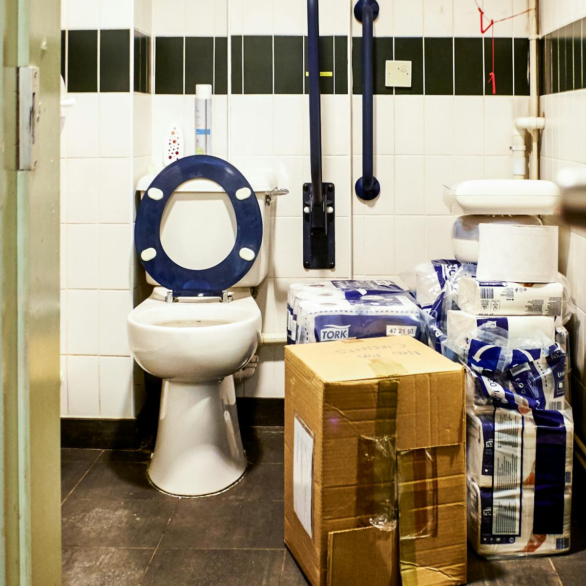 Photograph though a doorway into an accessible toilet. The floorspace next to the toilet is covered with a cardboard box and piles of packets of toilet paper.