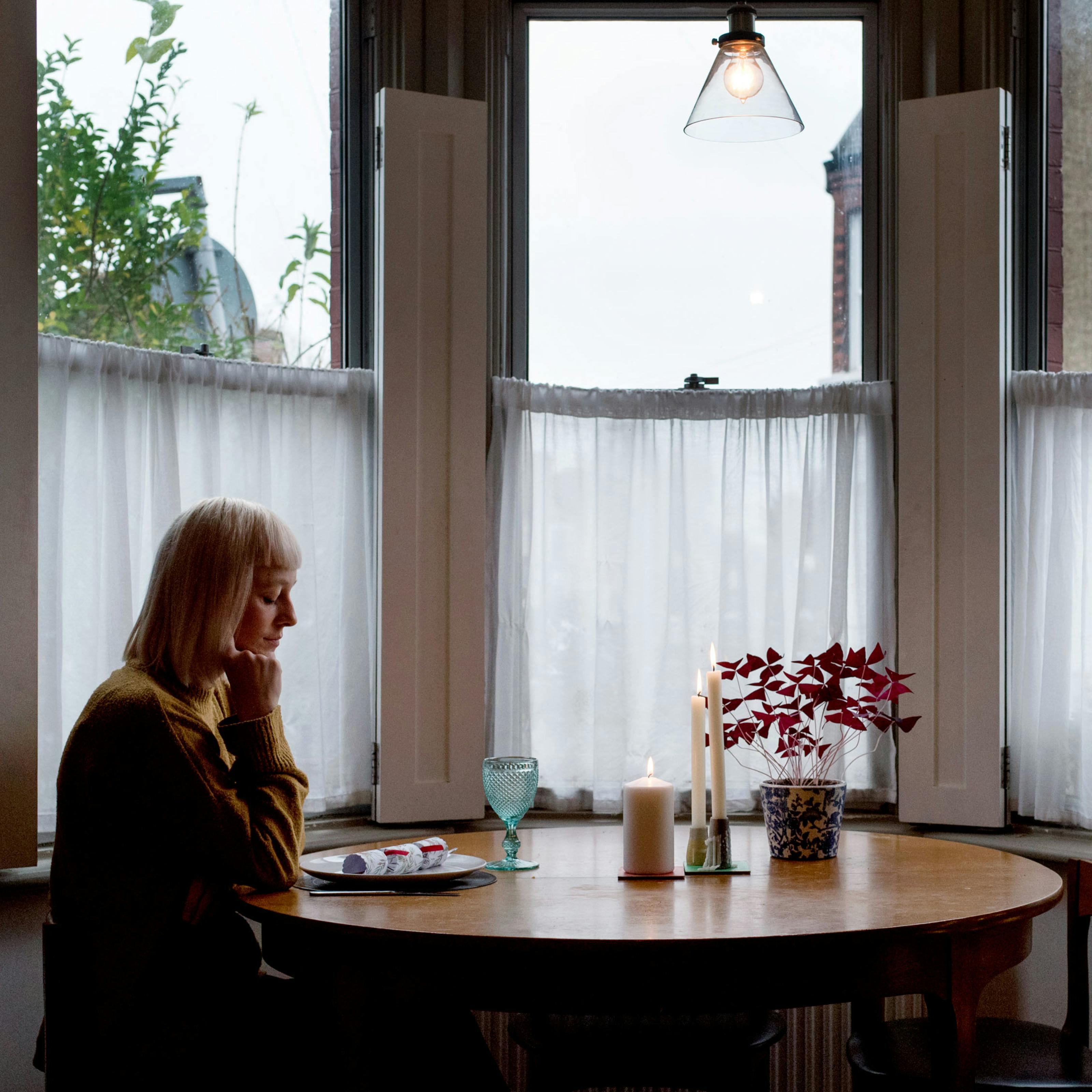 Photograph of a young woman sitting at a table laid for Christmas dinner for one. To the right hand side is a Christmas tree. Behind the woman is a bay window with net curtains covering the lower sashes.
