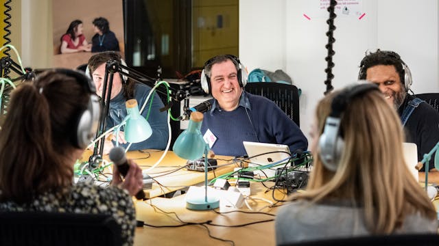 Photograph of a group of people sat around a table hosting a radio show. They are wearing headphones. The table is covered in wires from desk lamps, microphones and tablet devices. In the centre of the image one of the people is laughing.