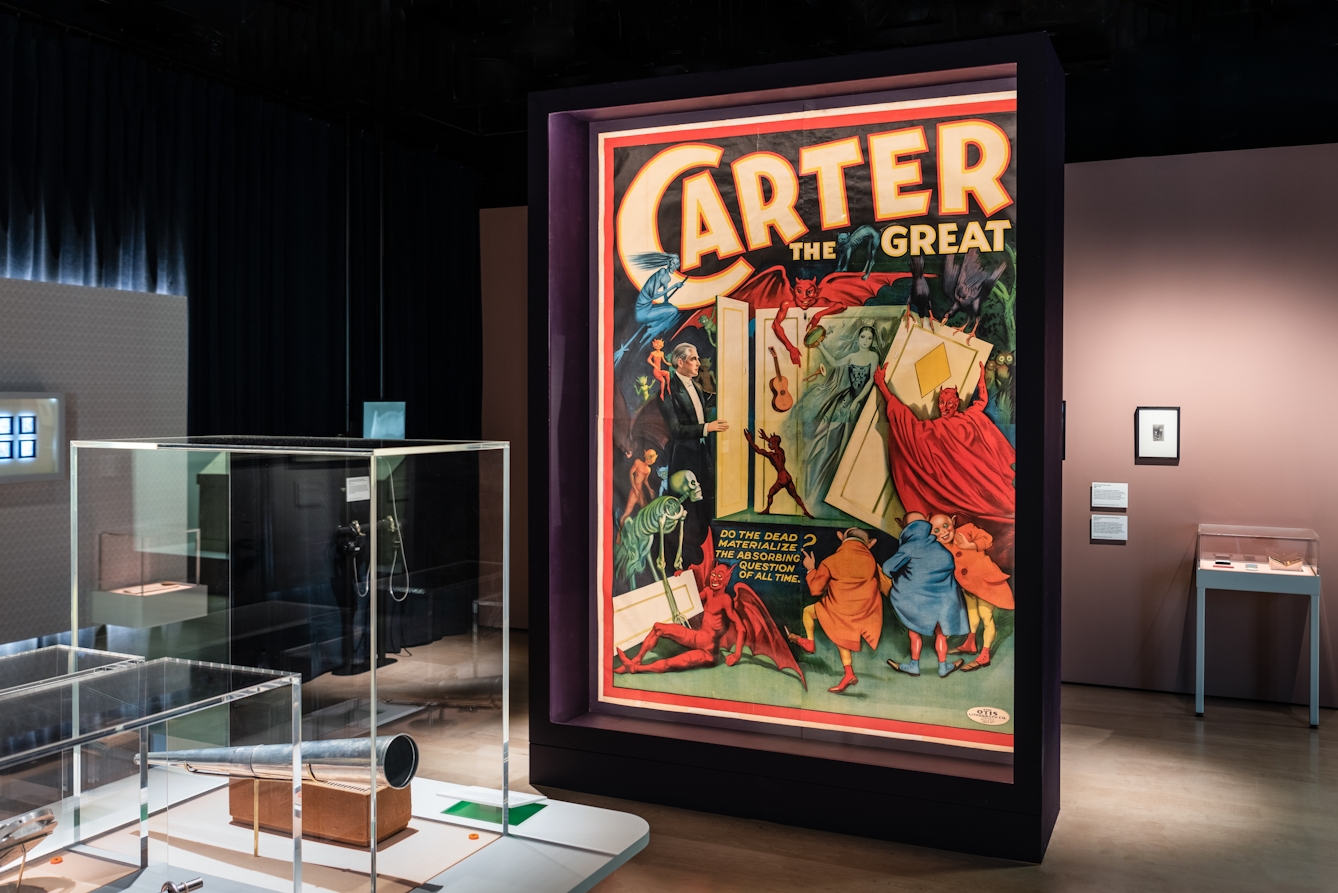 Photograph of a poster with text 'Carter the Great' with large colour graphic and subtext of 'Do the dead materialise? The absorbing question of all time', as part of the Smoke and Mirrors exhibition at Wellcome Collection.