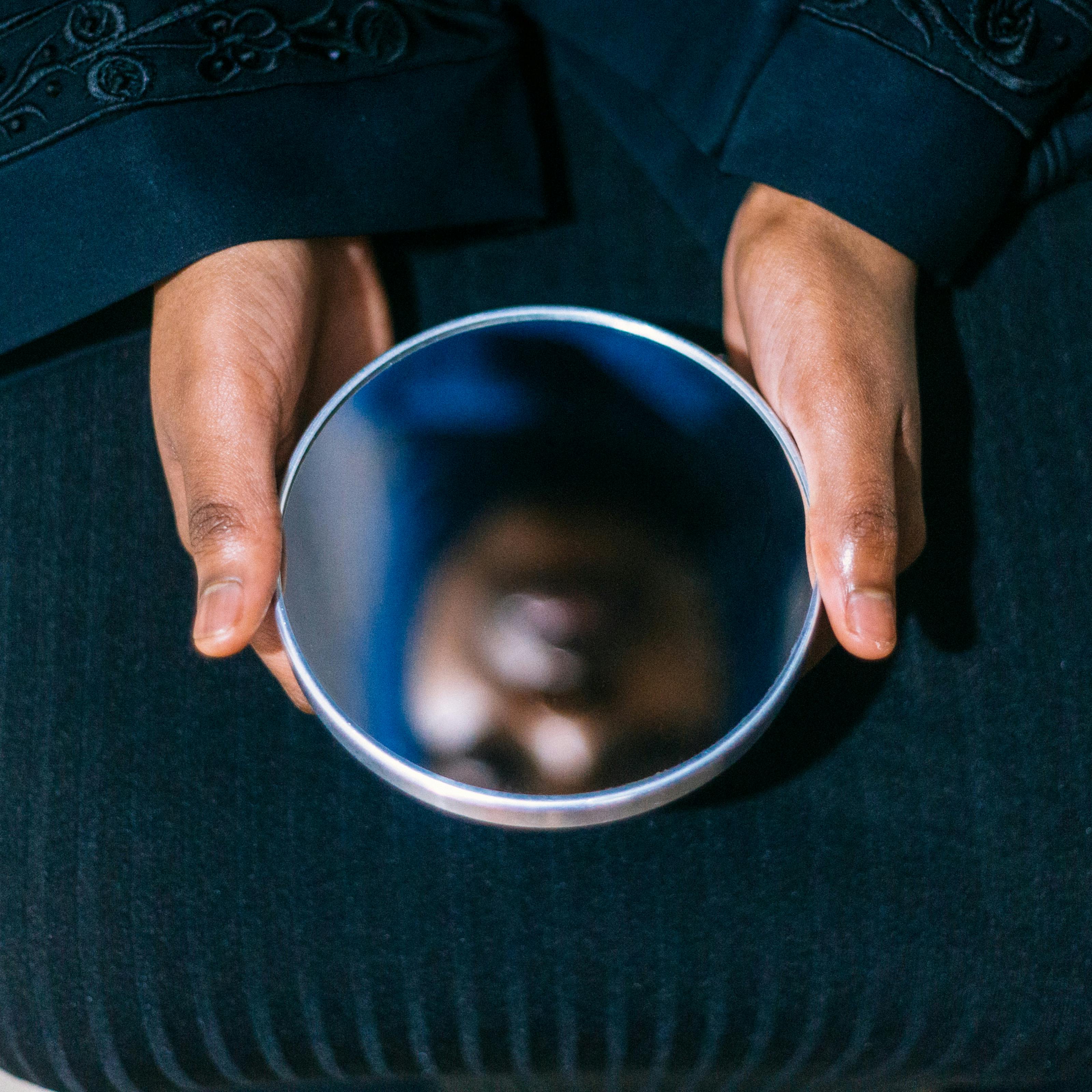 Photograph of a woman holding a circular mirror on her lap with both hands. In the reflection you can see her face, out of focus.
