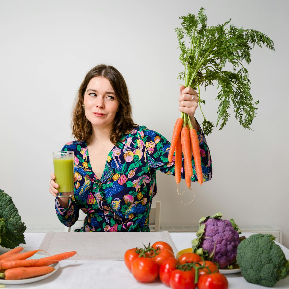 Photograph of writer Gwen Smith sat behind a table which is covered in vegetables, holding carrots in one hand and a green health drink in the other. She is looking sceptically at the vegetables.