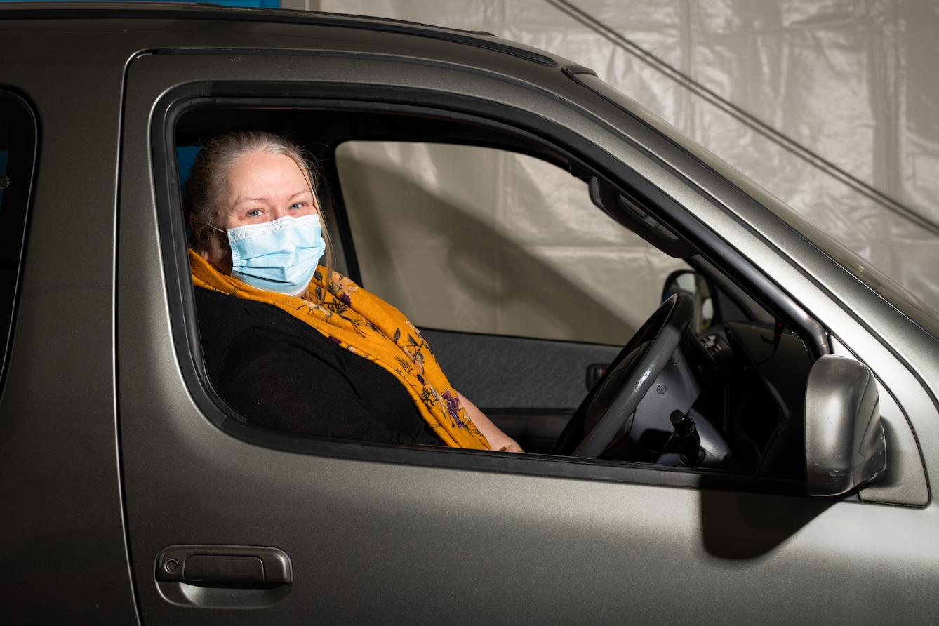 Photographic flash lit portrait through the open side window of a stationary car. The car is grey and the front of the car is facing to the right. Sat in the driver's seat wearing an orange floral shawl is a woman, looking to the camera. She is wearing a blue medical face covering. In the background is the out of focus inside of a white marquee.